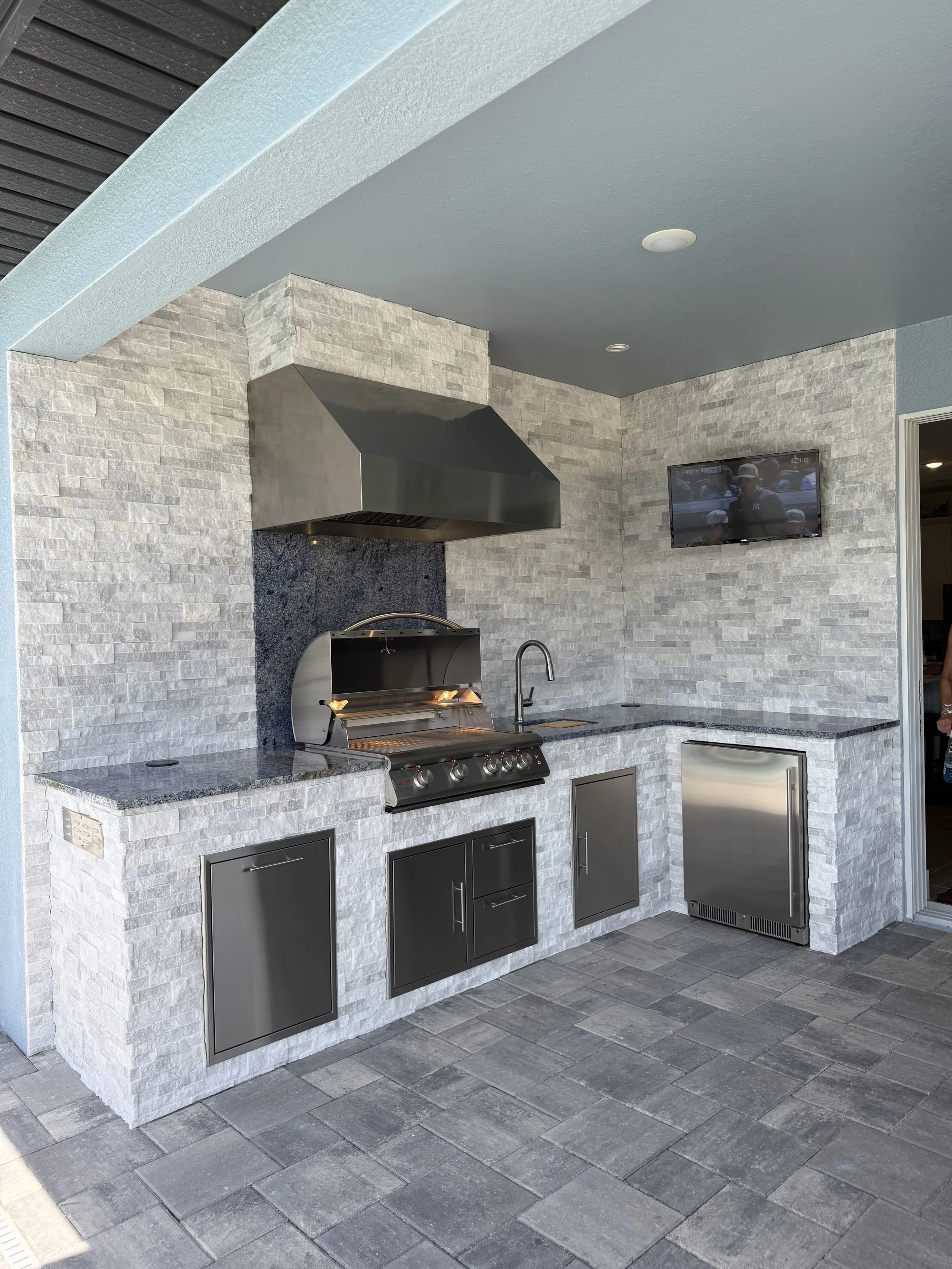Outdoor kitchen area with a stainless steel grill, gray cabinets, stone brick walls, granite countertops, and a small refrigerator. There is a wall-mounted TV and tiled flooring.