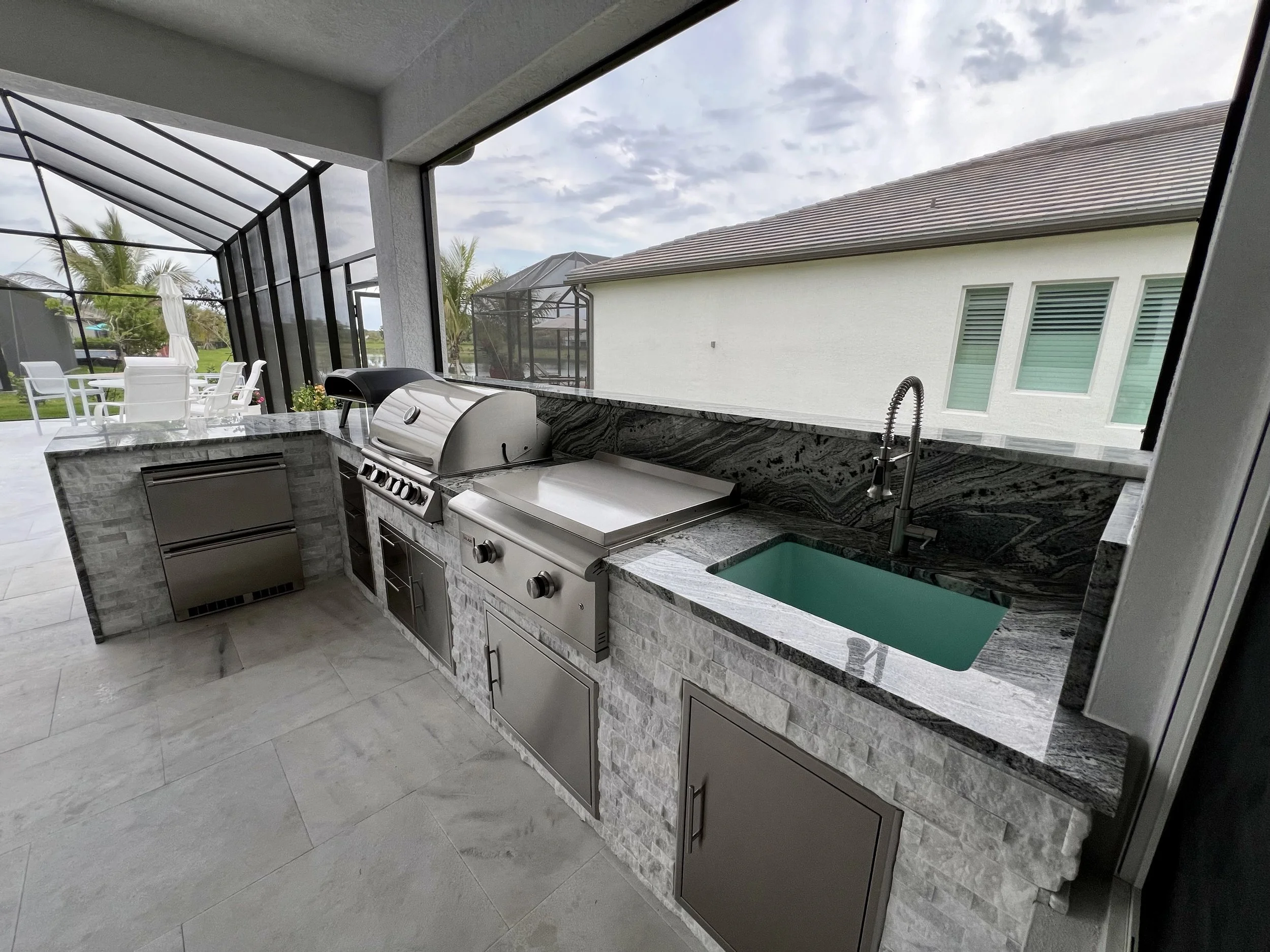 Outdoor kitchen with a stainless steel grill and sink, granite countertop, black-framed screen enclosure, patio furniture, and a view of a house and palm trees outside.