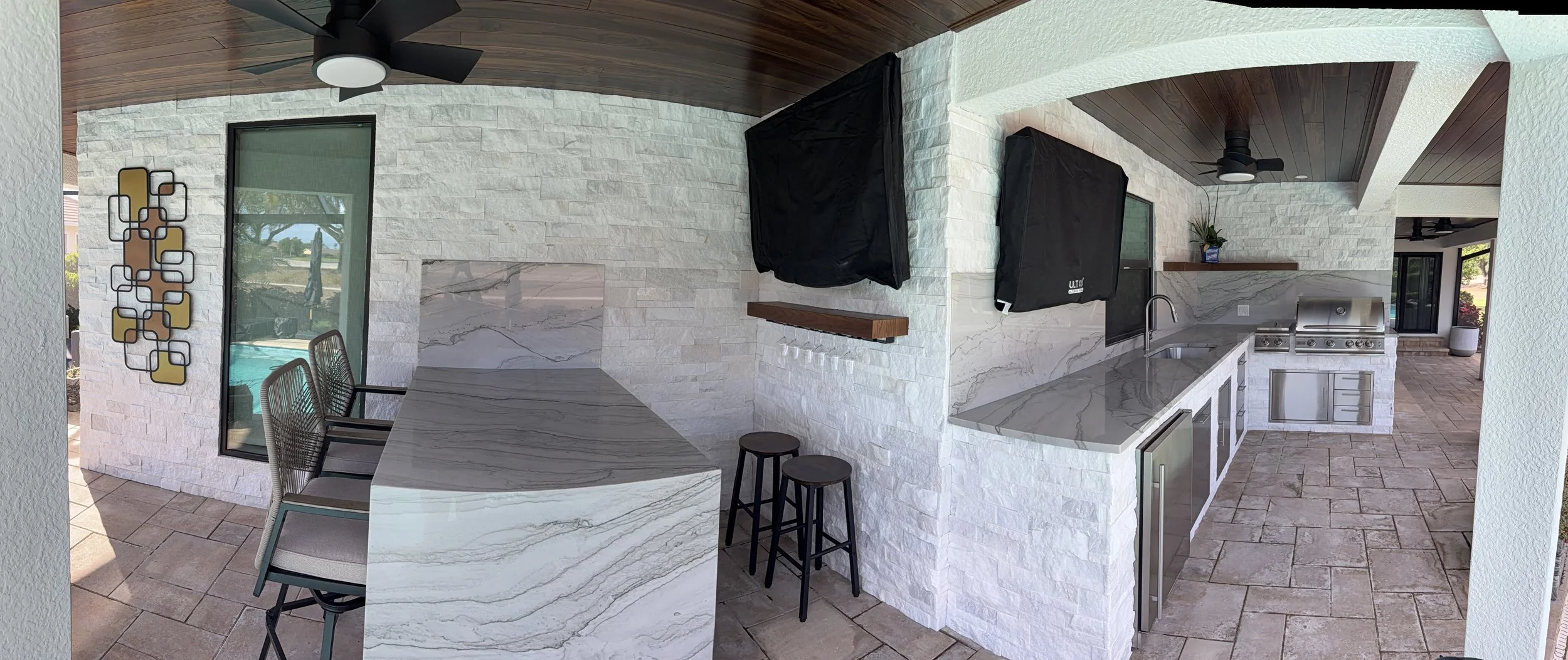 Covered outdoor kitchen area with a marble countertop, built-in grill, stainless steel mini-fridge, wall-mounted televisions, ceiling fans, and seating area with chairs, decorated with a modern wall art, stone walls, and potted plants.