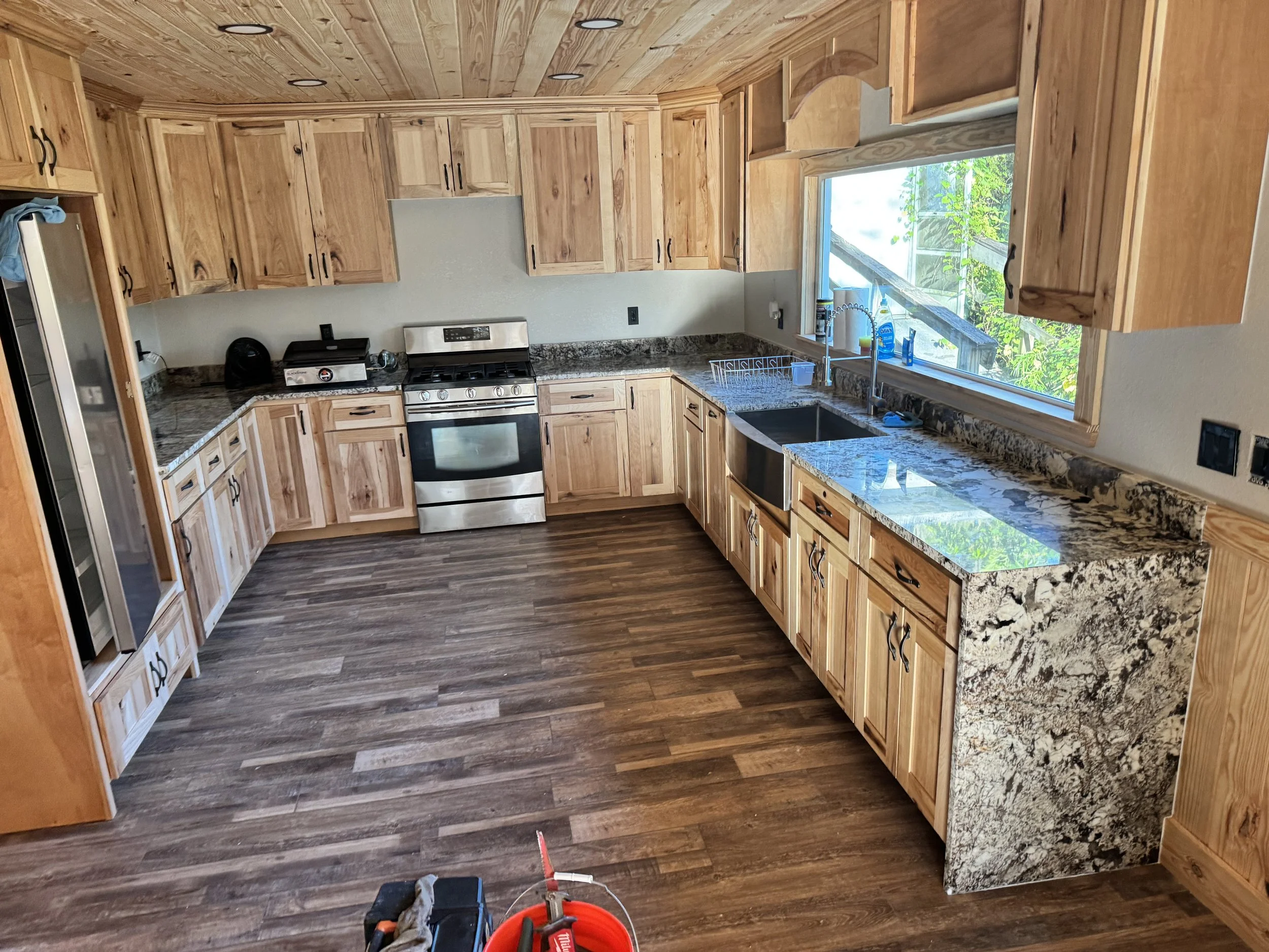 Kitchen with wooden cabinets, granite countertops, and a large window overlooking greenery. Contains stainless steel stove, dishwasher, and a sink. Construction tools and equipment on the floor.