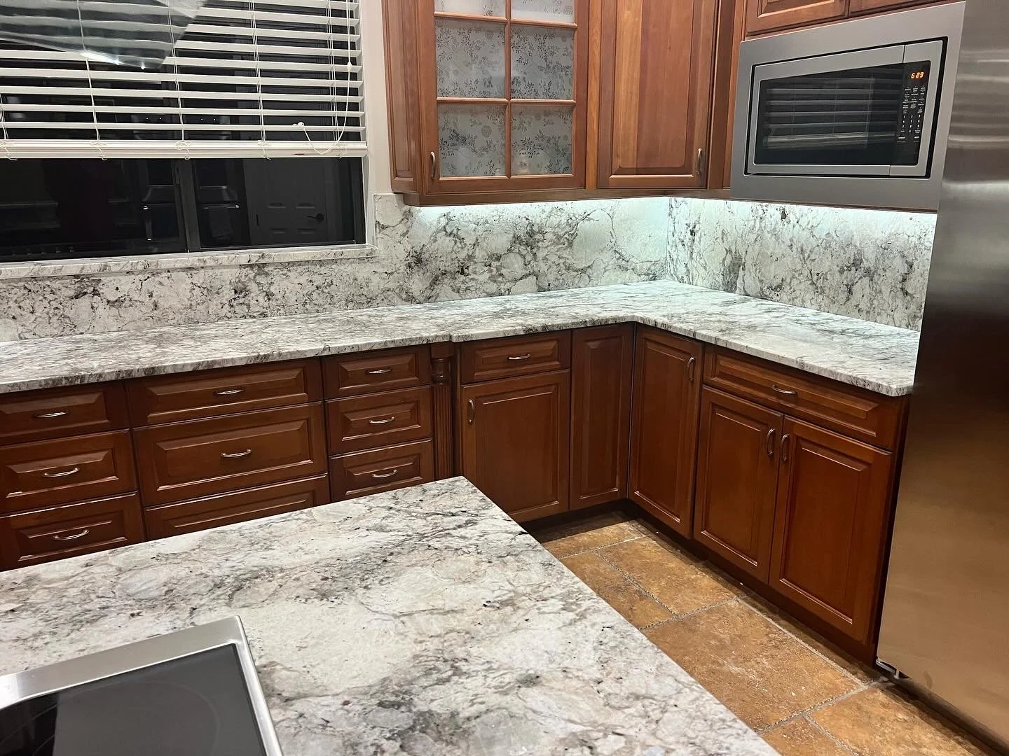 Kitchen with granite countertops, wooden cabinets, and stainless steel appliances, including a microwave. There is a window with blinds above the countertop.