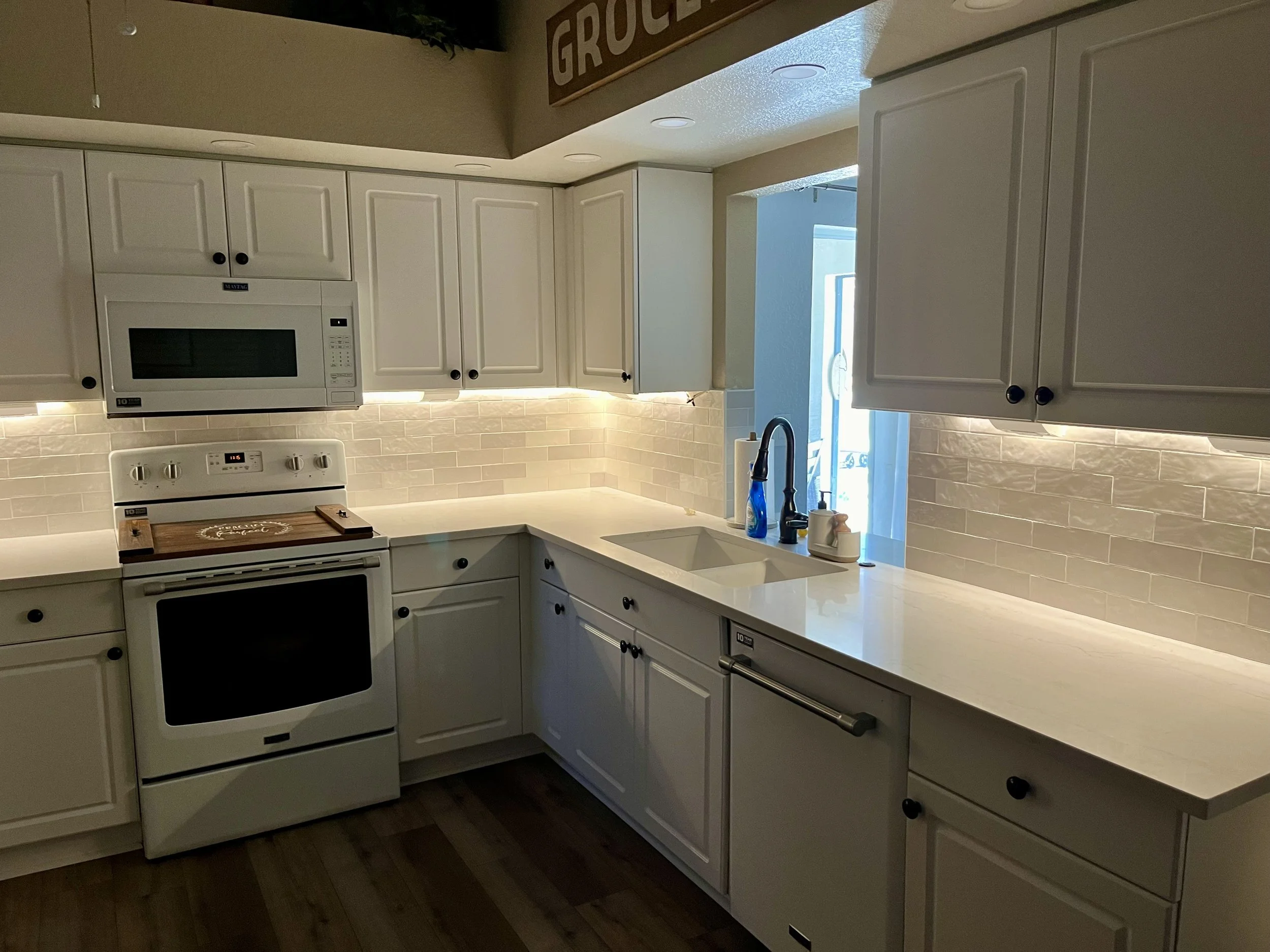 A kitchen with white cabinets, a stove, microwave, and a window near the sink with blue soap and paper towels, illuminated by ceiling lights and natural light.