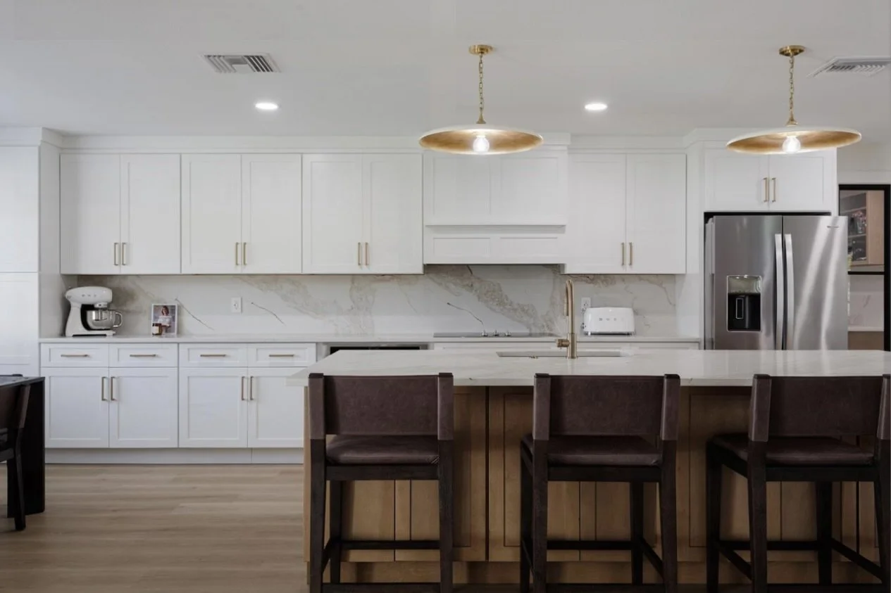 Modern kitchen with white cabinets, marble backsplash, and stainless steel refrigerator. Kitchen island with four brown chairs and two gold pendant lights.