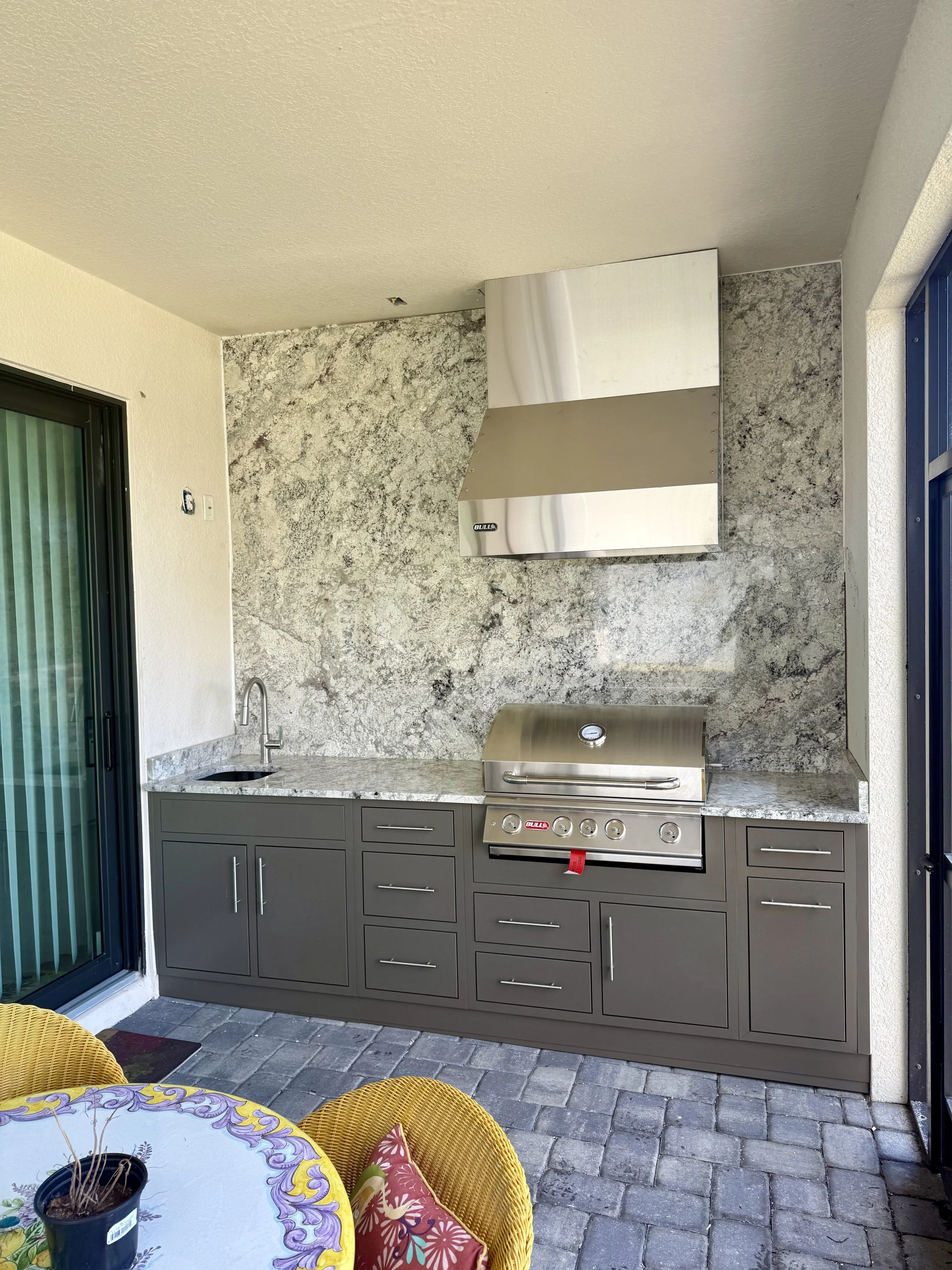 Outdoor kitchen area with a gray cabinet, built-in grill, stainless steel range hood, granite countertop, a small sink, and a sliding glass door with vertical blinds. There is a patio with stone pavers and a round table with yellow chairs.