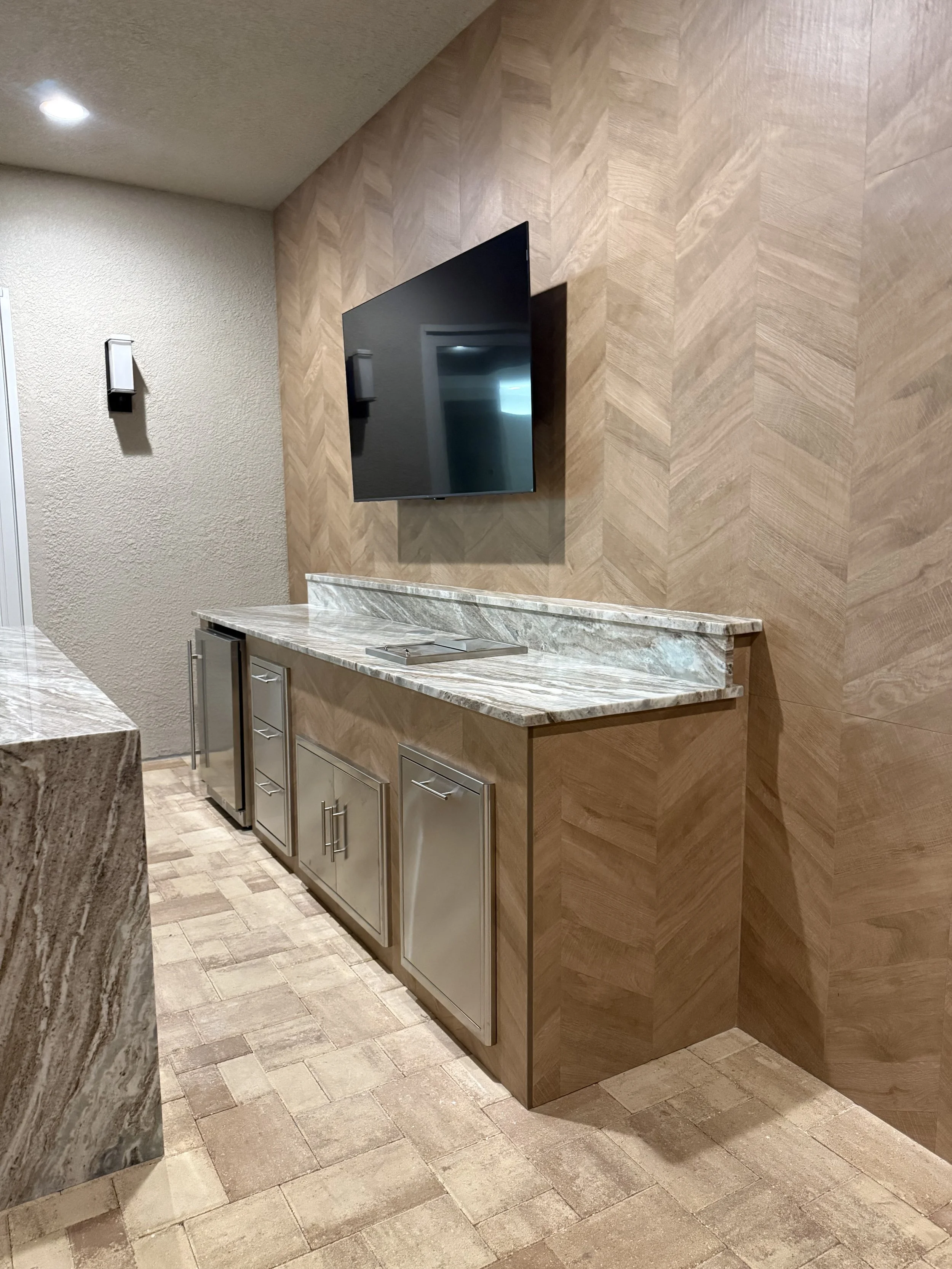 A modern interior wall with wood paneling, a flat-screen TV mounted on it, a marble countertop, and a stainless steel cabinet below the counter.