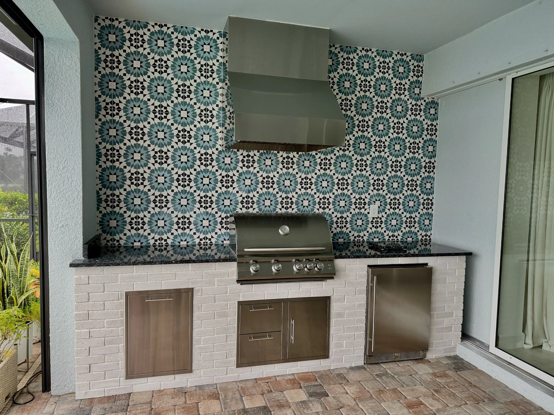 Outdoor kitchen with a built-in stainless steel grill, black granite countertop, white brick base, and Moroccan-style patterned tile backsplash. There is a sliding glass door on the right and a patio area with brick flooring.