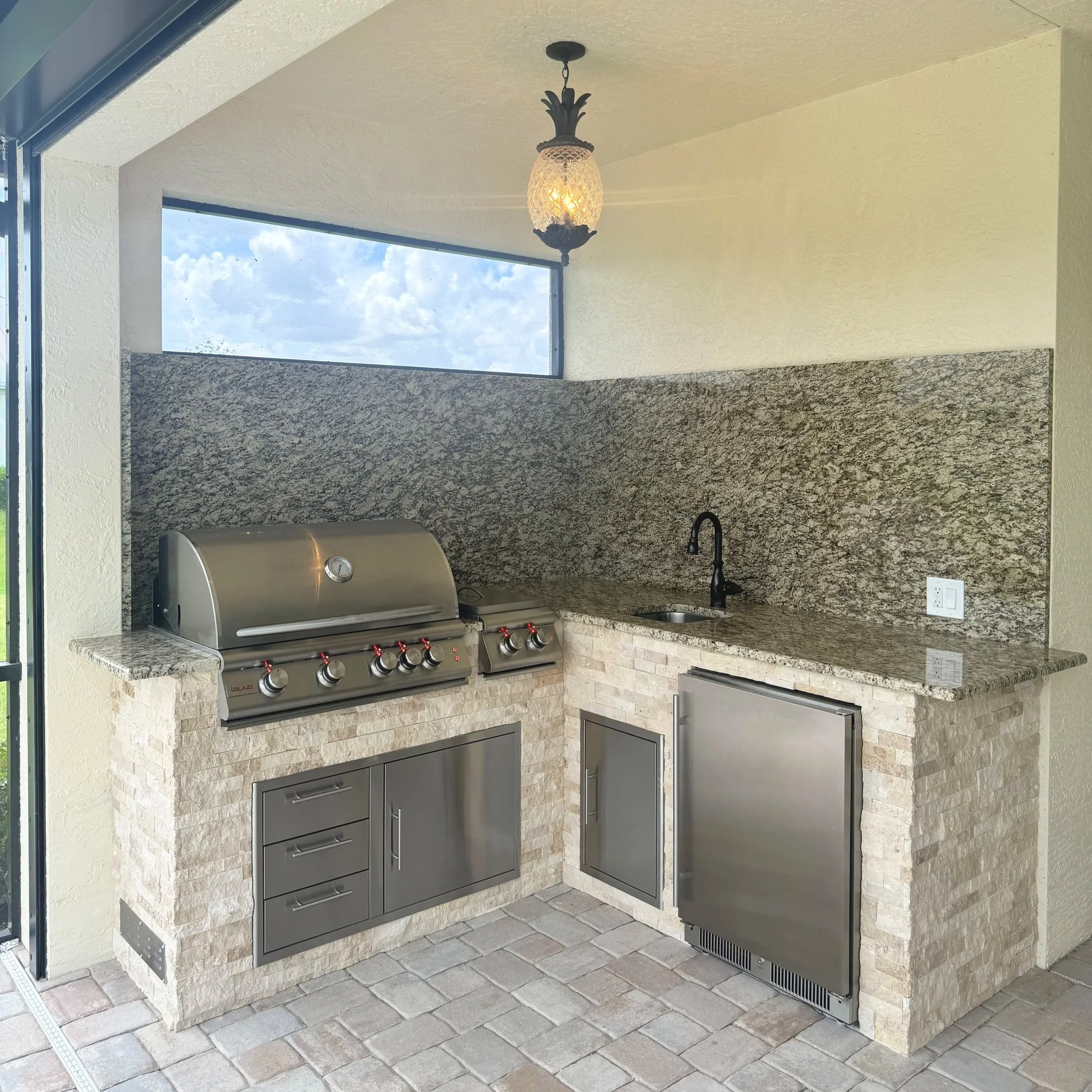 Outdoor kitchen with stainless steel grill, granite countertops, a stone wall, and a hanging lantern with cloudy sky visible through a window.