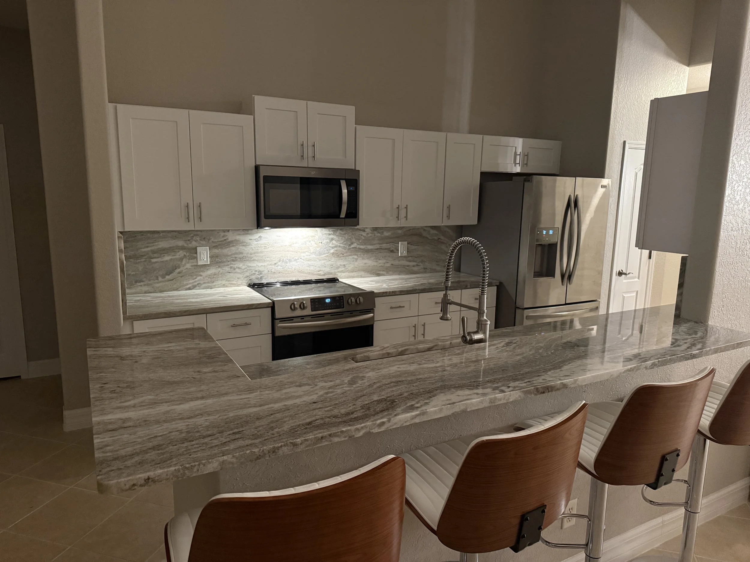 Modern kitchen with white cabinetry, stainless steel appliances including a microwave and refrigerator, marble countertops, and a kitchen island with bar stools.