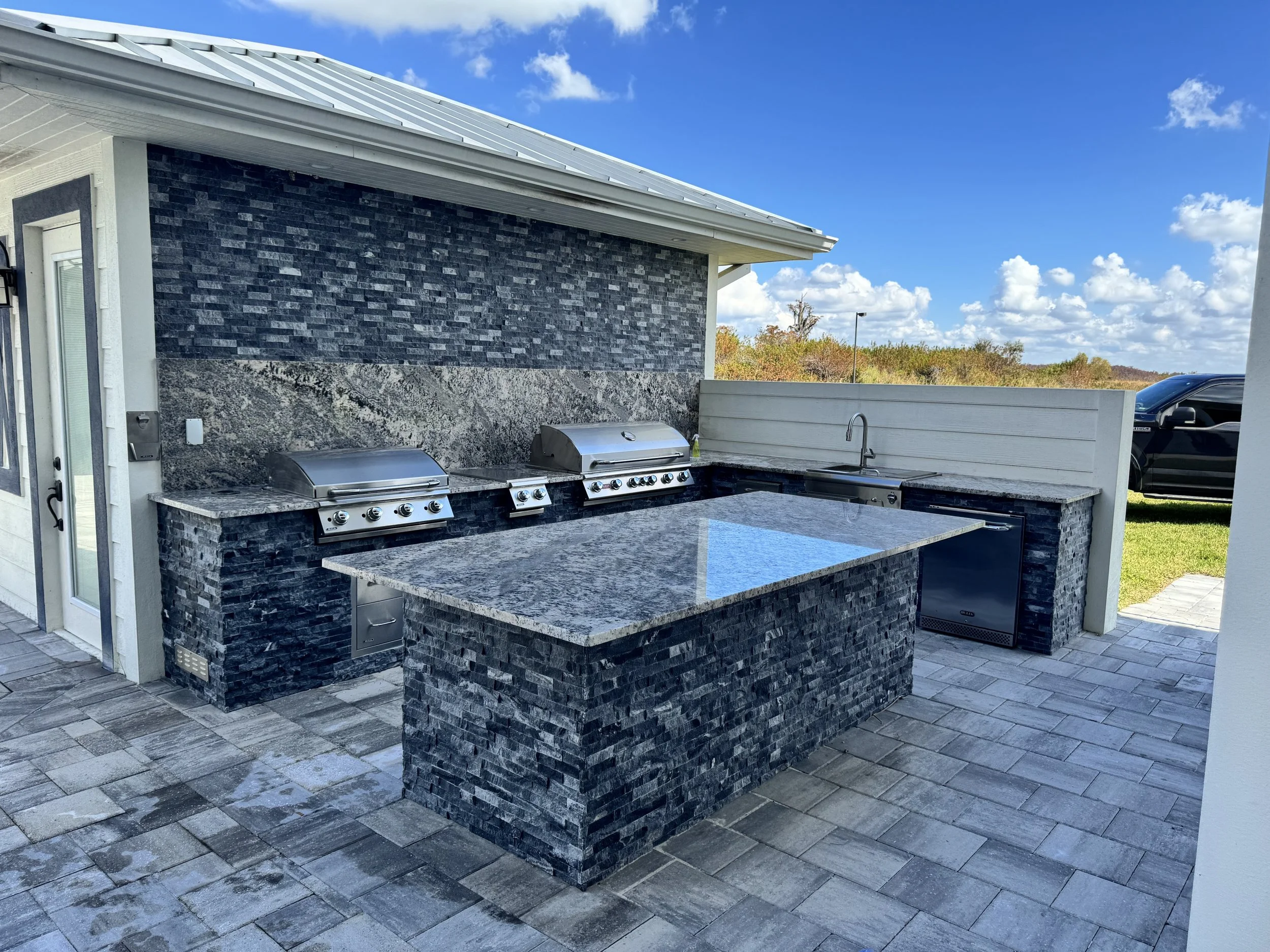 Outdoor kitchen area with two stainless steel grills, a stone countertop, a sink, and a textured stone and tile backsplash, under a metal roof with a blue sky and clouds in the background.