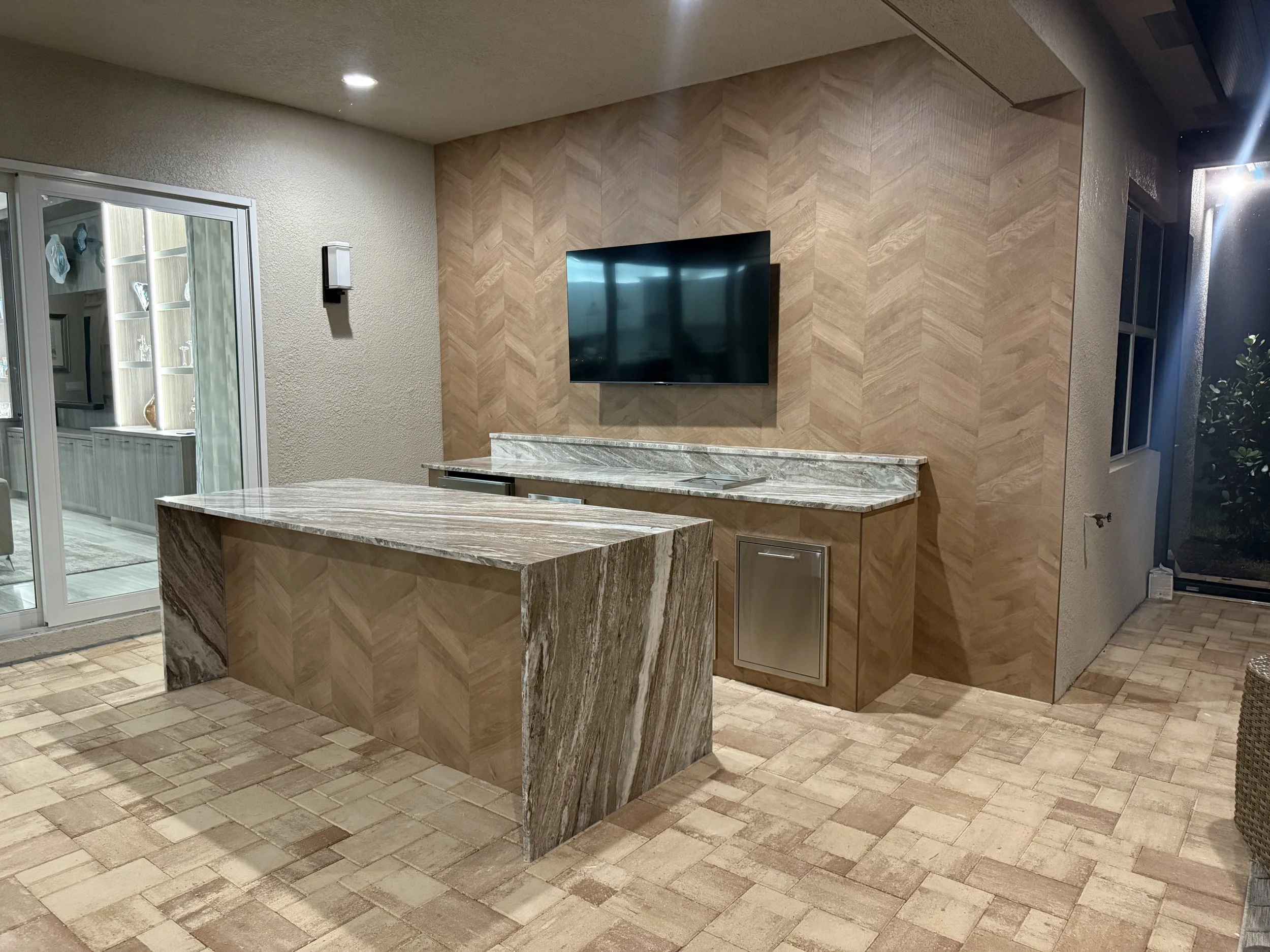 Modern living room with a wall-mounted flat-screen TV, patterned wood wall, marble countertop, sliding glass door, brick floor, and partially visible sitting area.