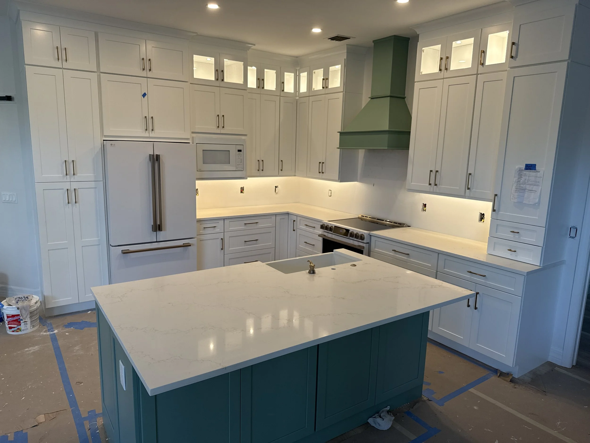 Kitchen with white cabinets, a white island with countertop, and a green range hood, under construction with painter's tape and tools visible.