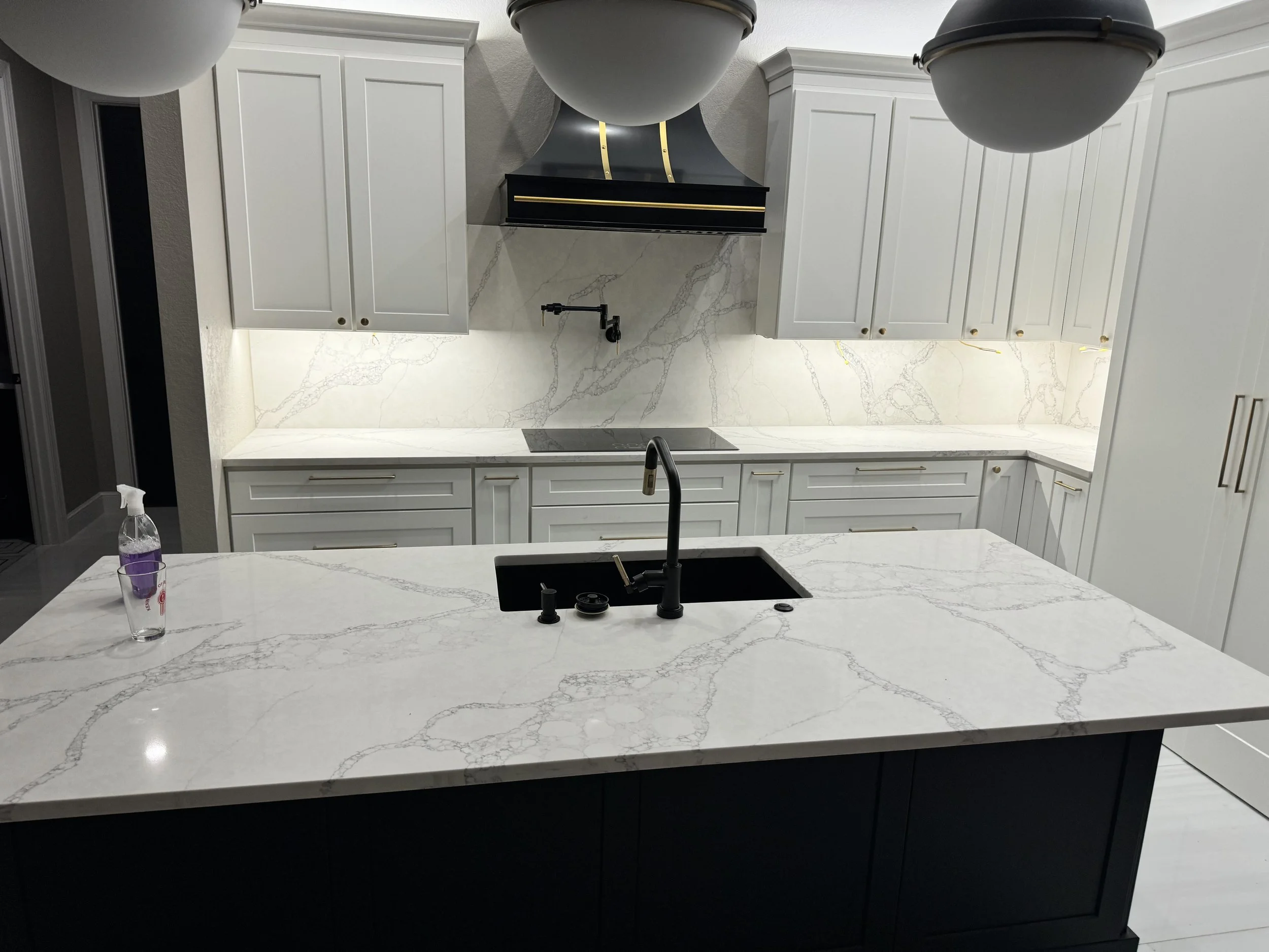 Modern kitchen with white marble countertops and backsplash, white cabinets, black range hood, black faucet, and a bottle of purple cleaning spray on the counter.