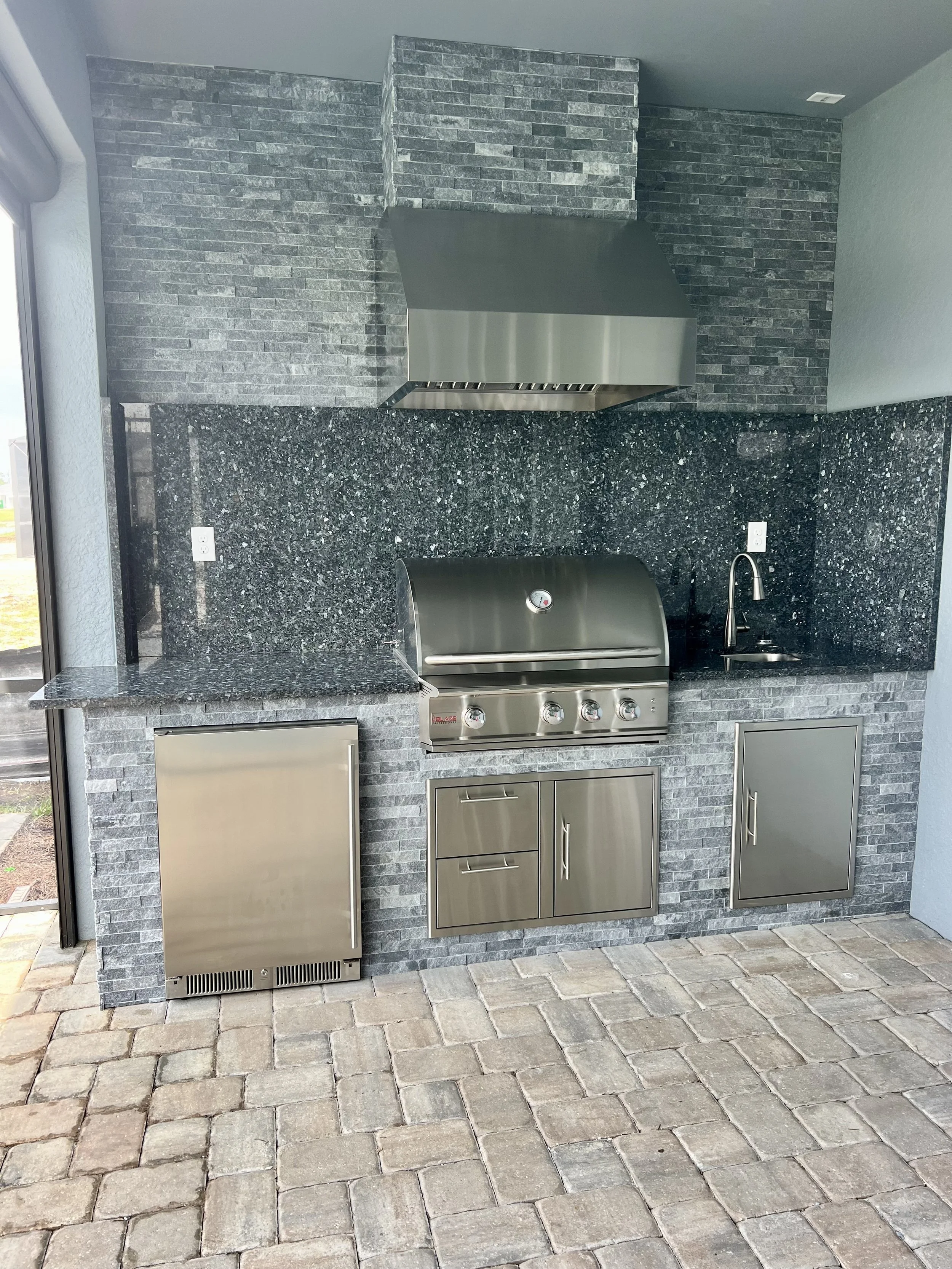 Outdoor kitchen with gray brick wall, stainless steel grill, cabinetry, black granite countertop, and stone pavement floor.