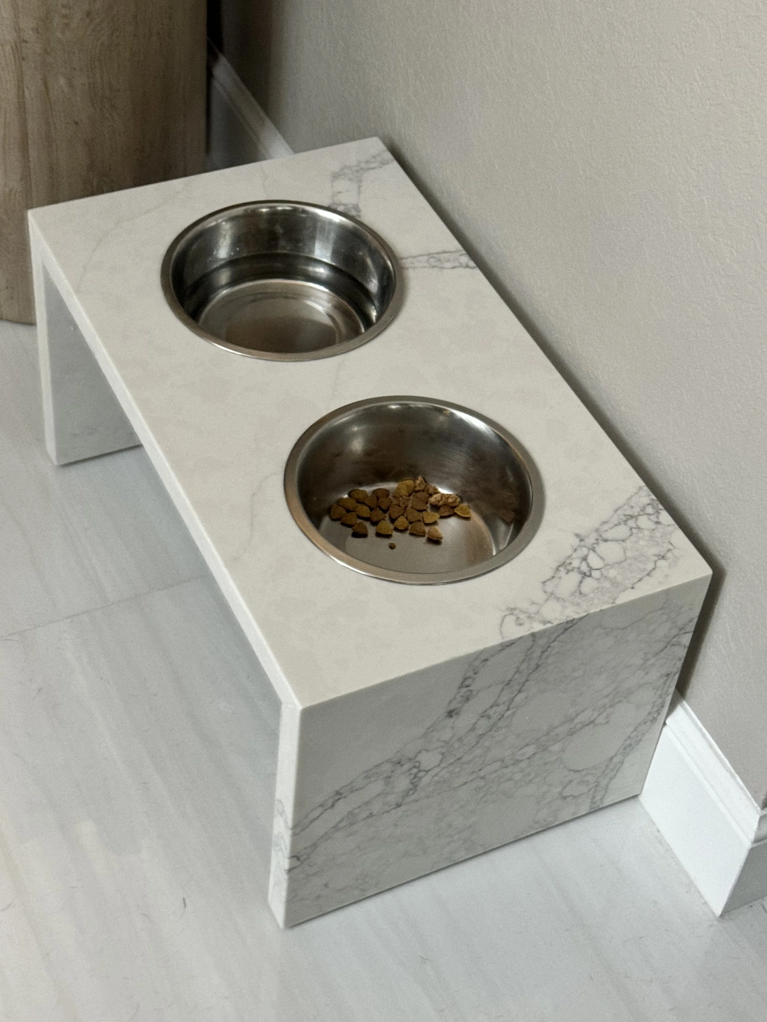 A white marble pet feeding station with two stainless steel bowls, one containing dry pet food, positioned against a wall in a room with a wooden door.