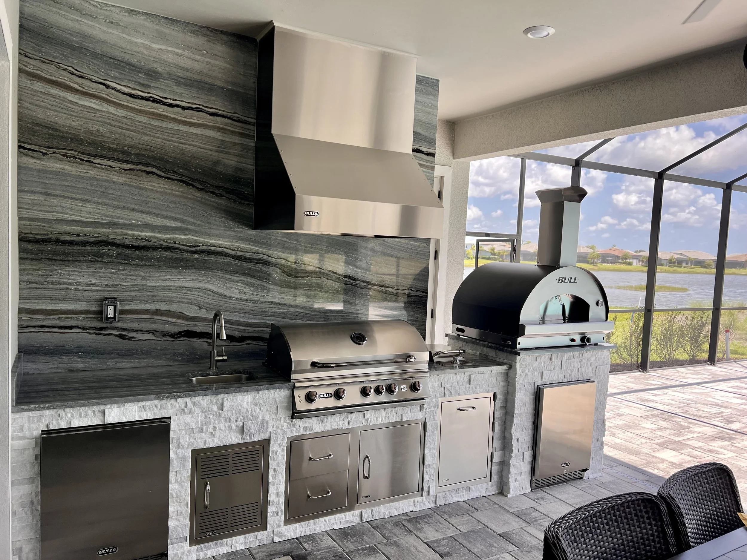 Outdoor kitchen with a grey stone countertop, a stainless steel grill and hood, black vents, and a view of houses and water through large screened windows.