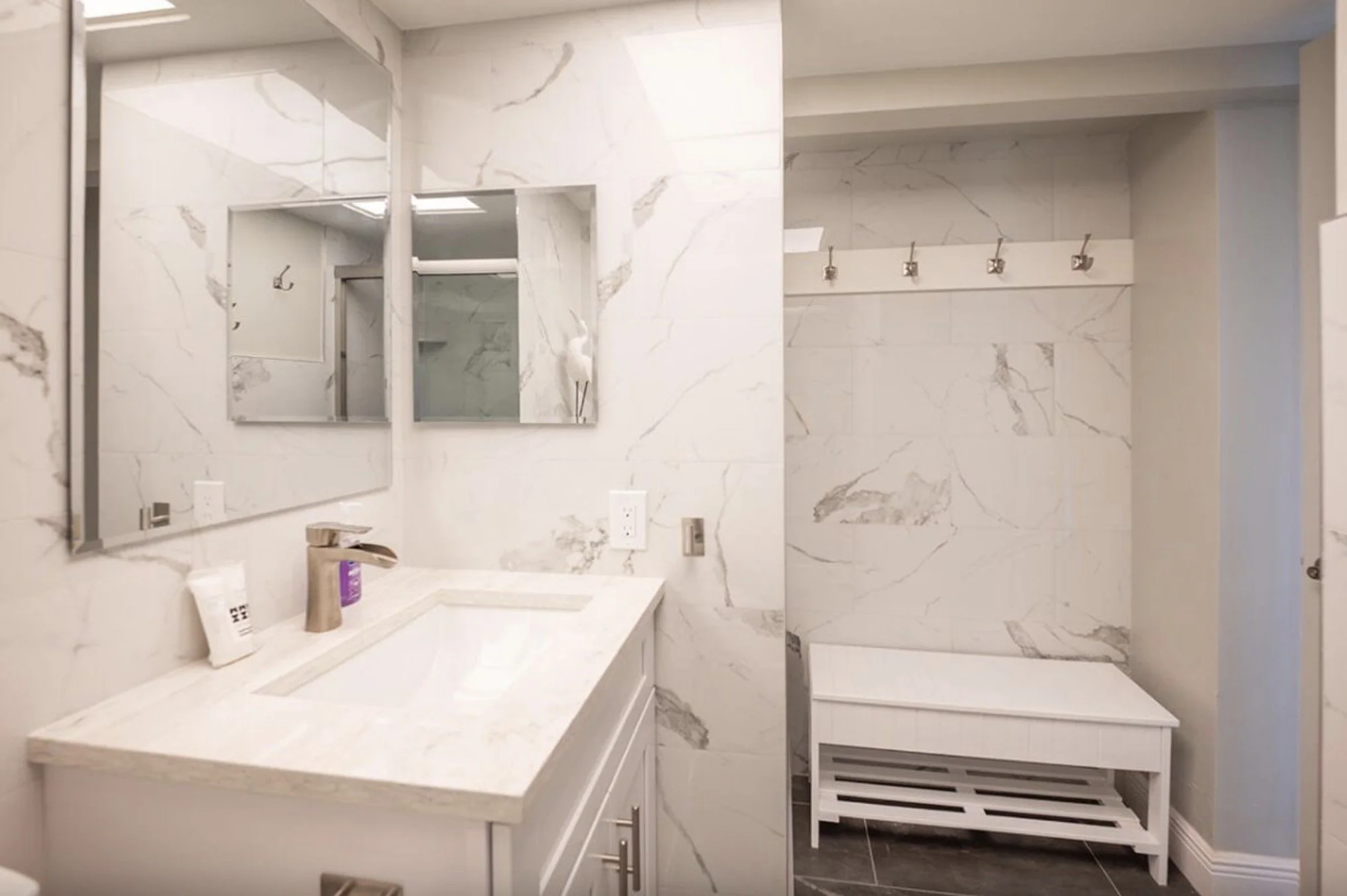 Bathroom with white marble walls and a white marble countertop, featuring a modern faucet, and a large mirror. There are hooks on the wall for hanging towels and a vent below a bench or storage area.