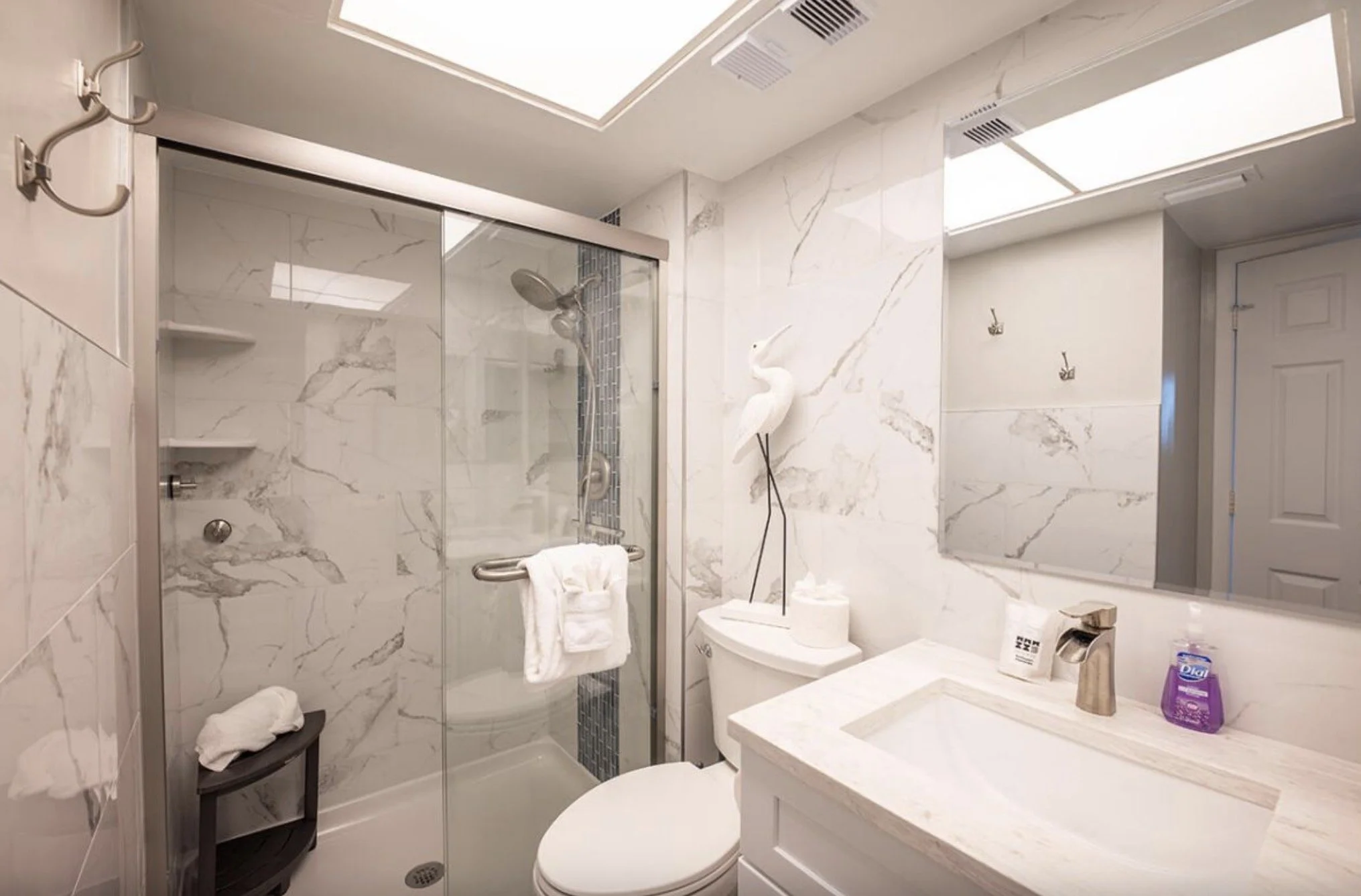 A modern bathroom with marble walls and a glass shower enclosure. There is a white toilet, a marble countertop with a sink, and a decorative white heron statue. Items on the counter include a purple container of disinfectant spray. The bathroom has c