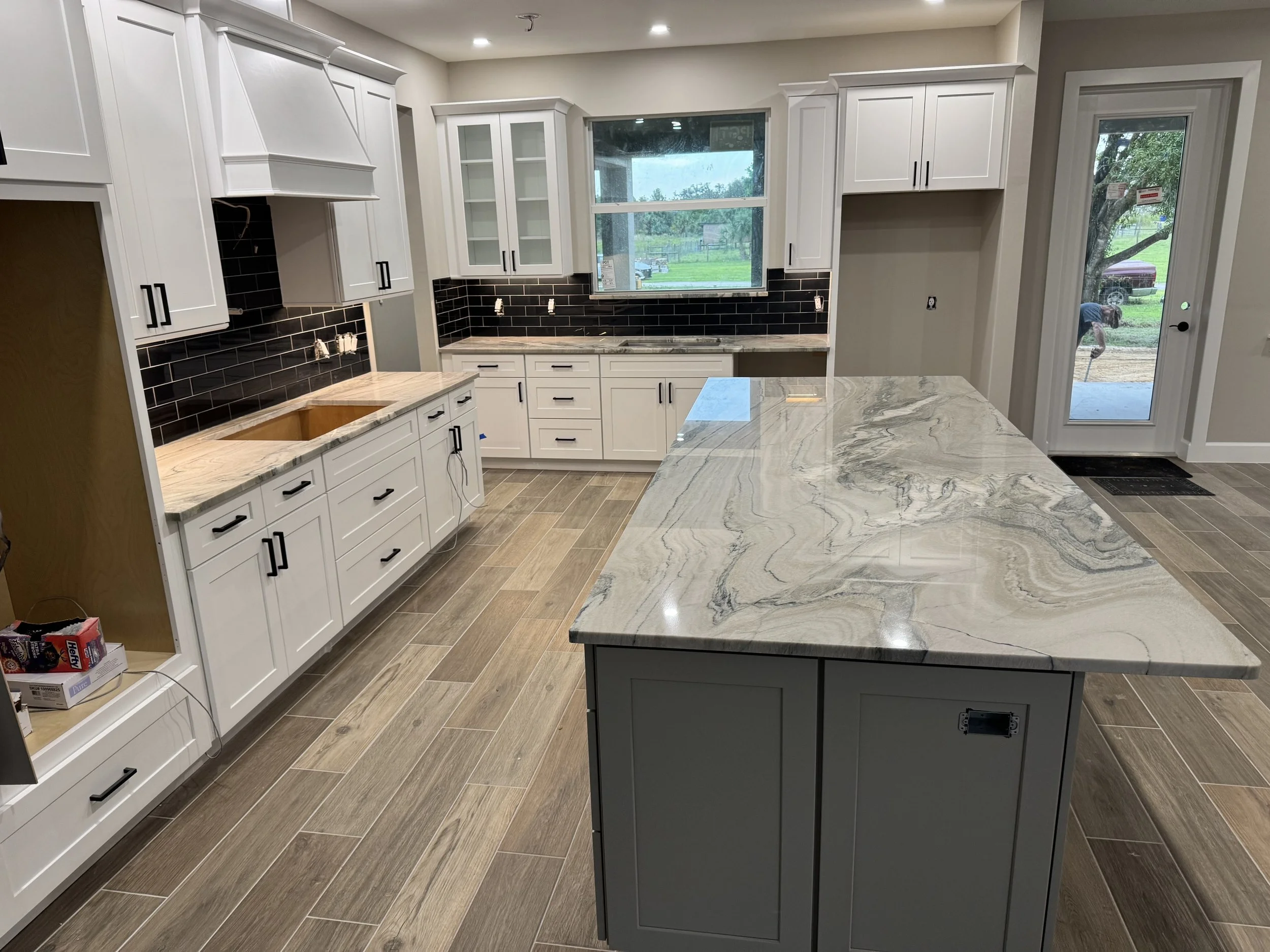 A kitchen under construction with white cabinets, black hardware, black subway tile backsplash, gray marble countertops, and a large window showing a backyard with green grass and trees.