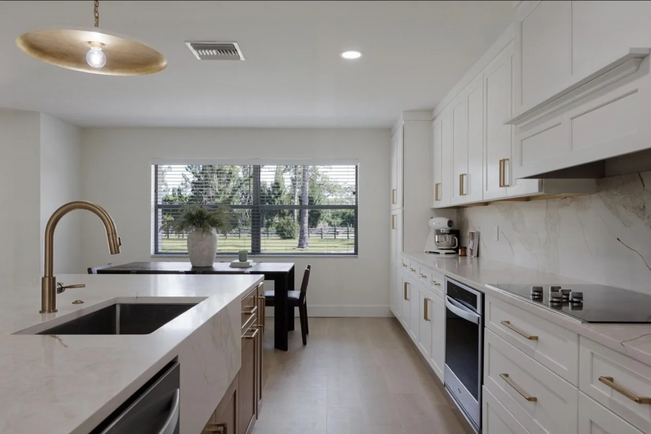 Modern kitchen with white cabinets, marble countertops, and gold handles. Includes an oven, stovetop, coffee maker, and a large window overlooking a green outdoor area. Kitchen island has a gold-colored faucet and a black sink.