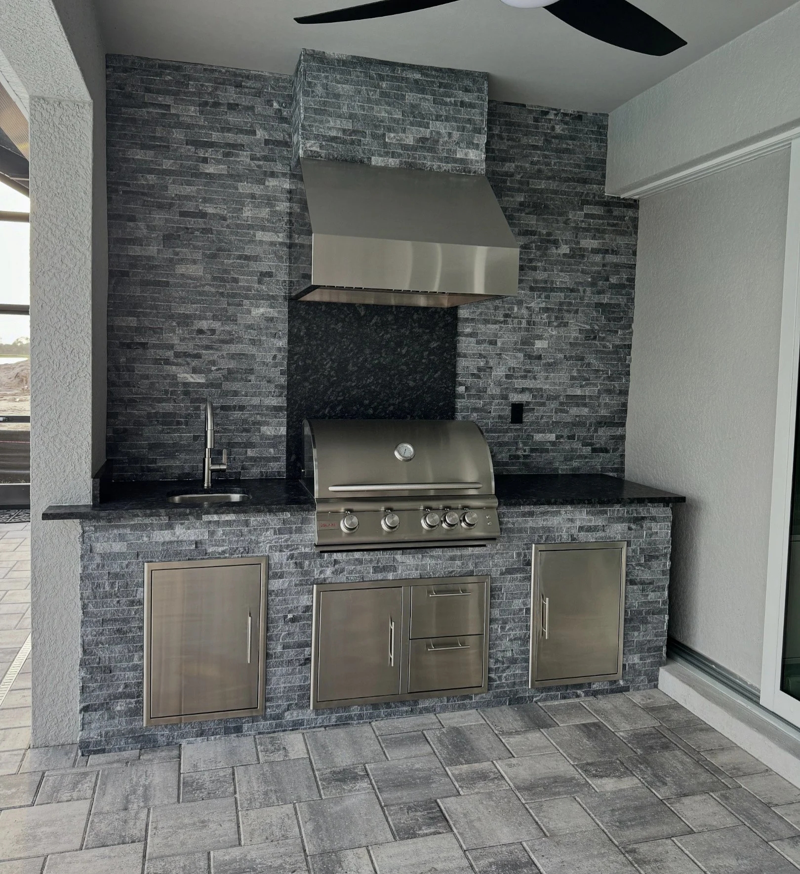 Outdoor kitchen area with a stainless steel grill, black granite countertop, black tile backsplash, and wall-mounted cabinets in a modern style.