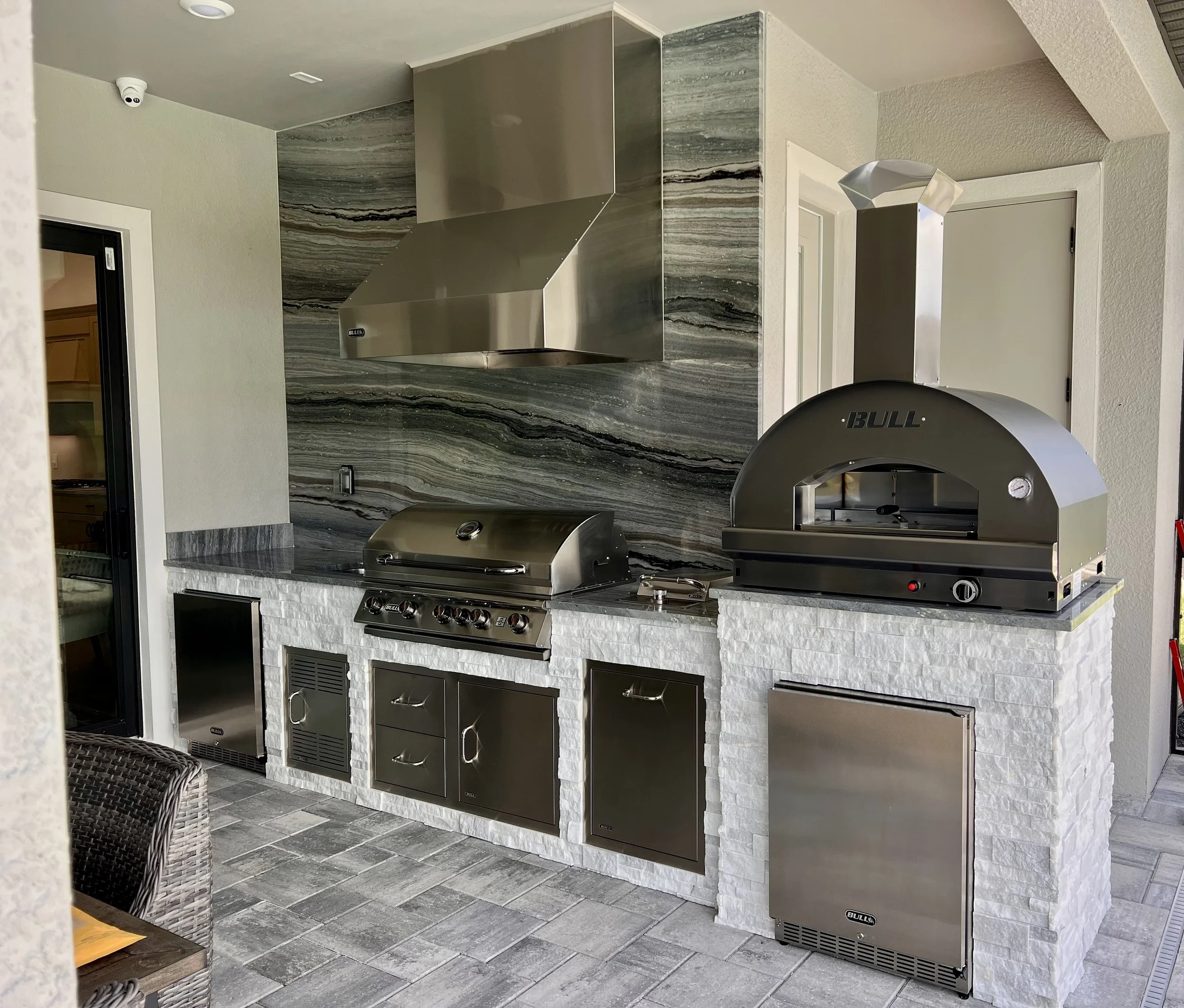 Outdoor kitchen area with a stainless steel grill, a pizza oven, and a marble-like stone backsplash. The area features gray tiles on the floor and white textured walls, with a door to the left and a ventilation hood above the grill.
