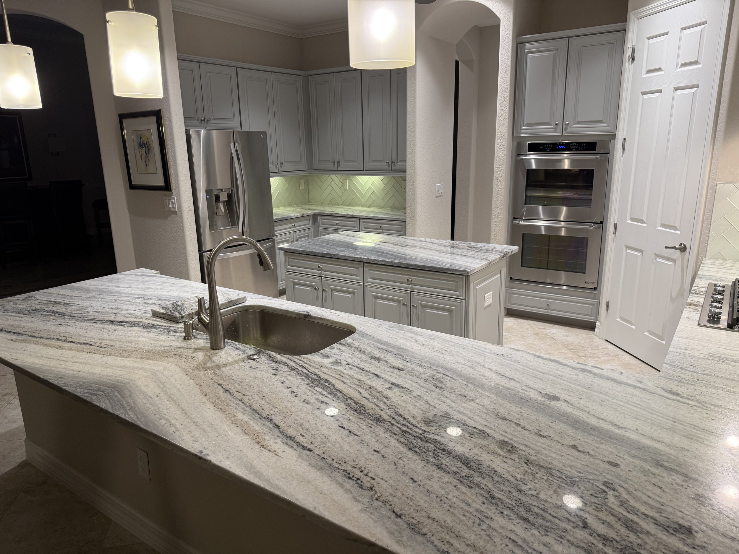 Kitchen with gray cabinets, stainless steel refrigerator, double oven, and a large granite island with a sink.