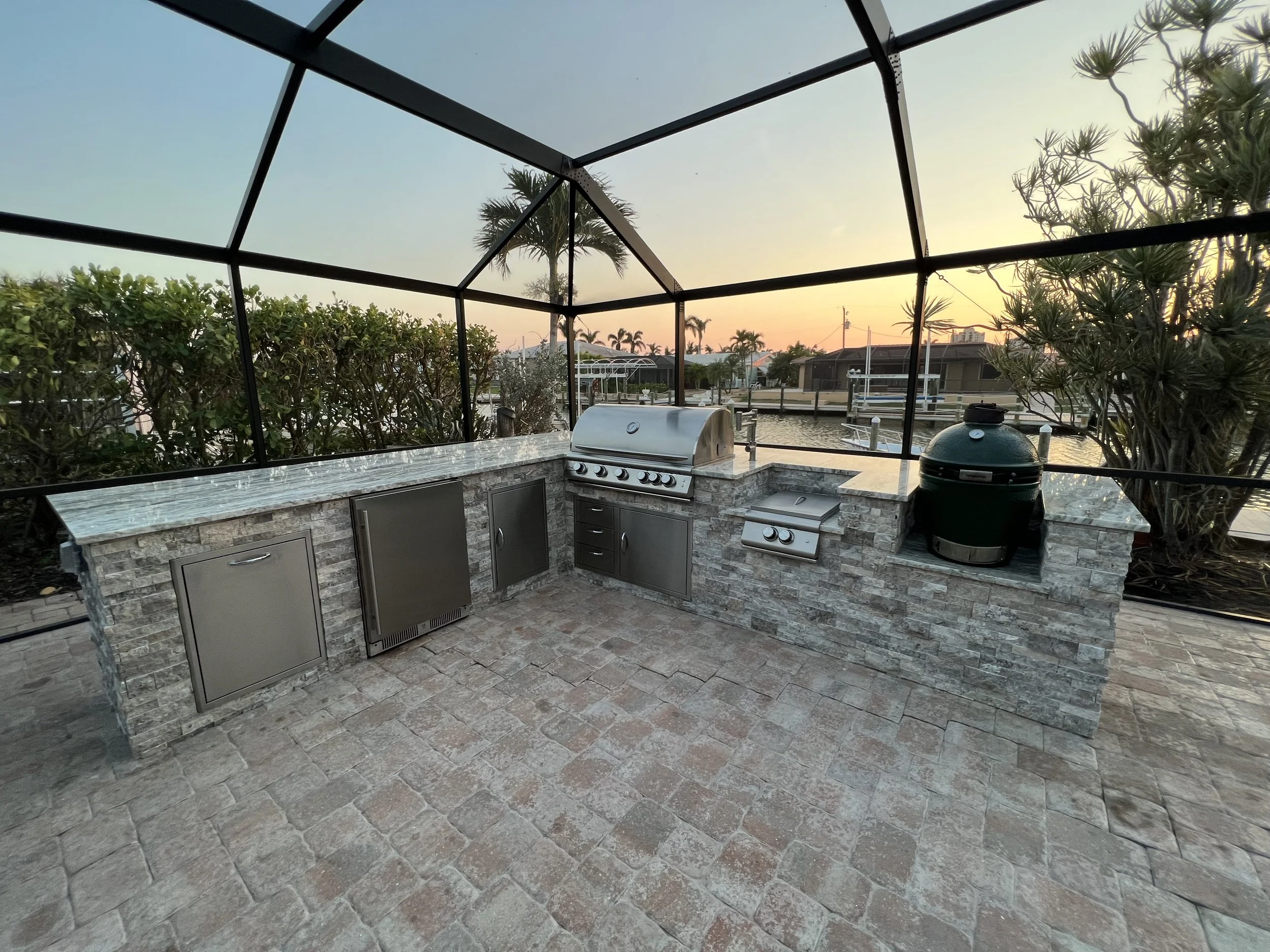 Outdoor kitchen area with a built-in grill, counter space, and a green ceramic grill, overlooking a canal with boats and homes at sunset.