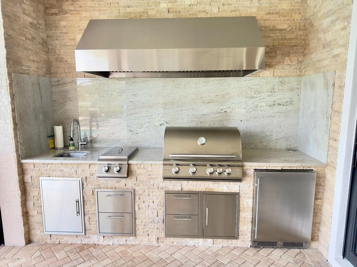 Outdoor kitchen with brick walls, granite counter, stainless steel grill, range, small sink, and cabinet storage.