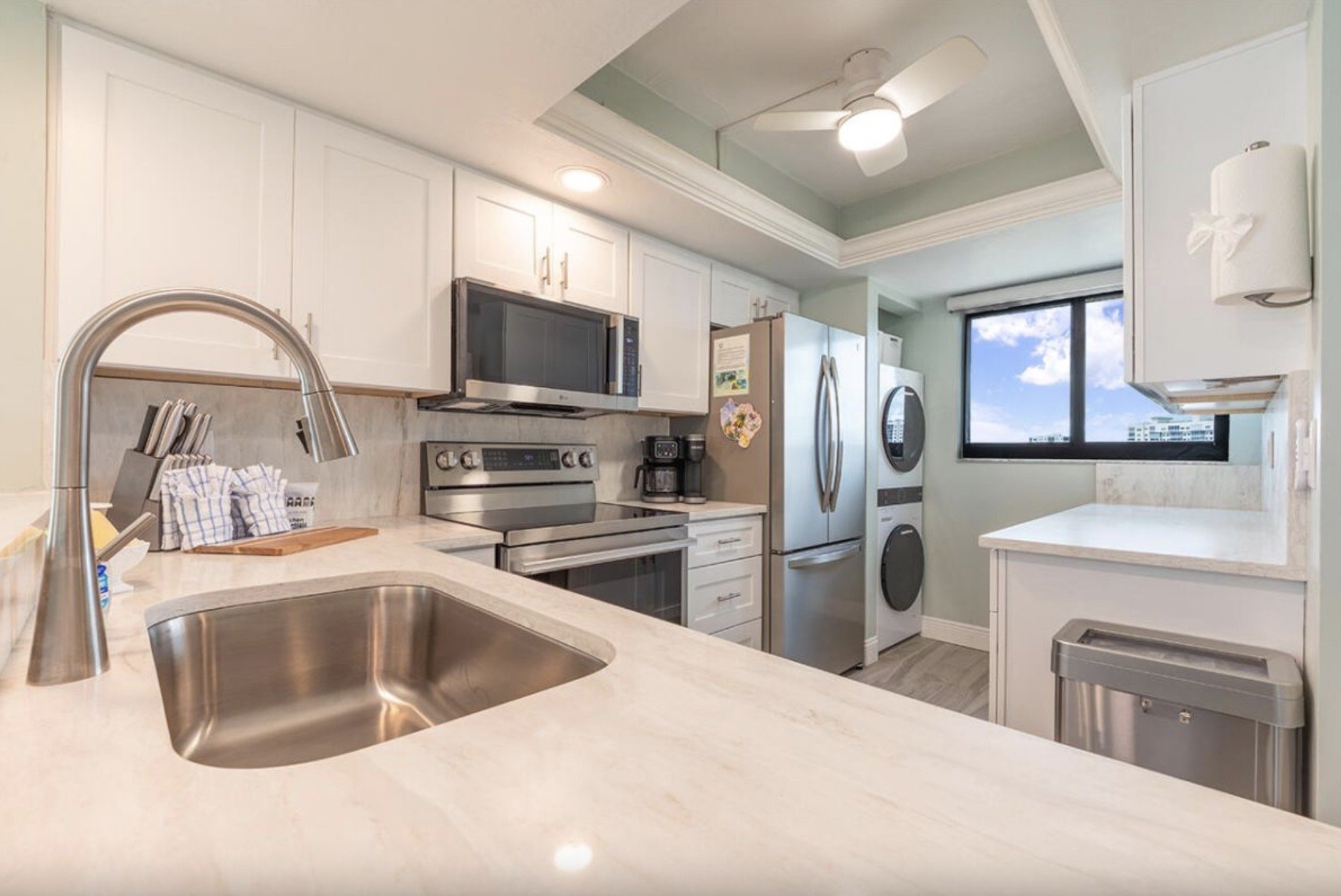 Modern kitchen with white cabinets, stainless steel appliances, a window with a city view, and a white countertop with a sink.