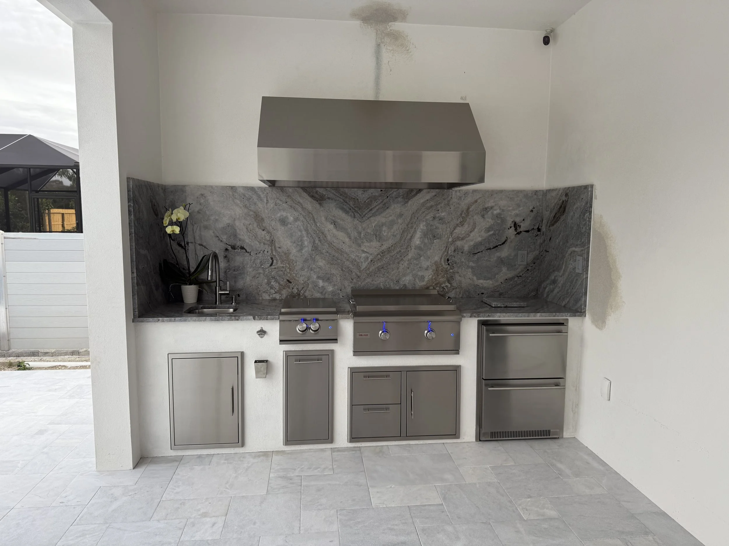 Modern outdoor kitchen with marble countertops, stainless steel appliances, and cabinets, featuring a sink, orchid plant, and textured backsplash, with an adjoining patio area.