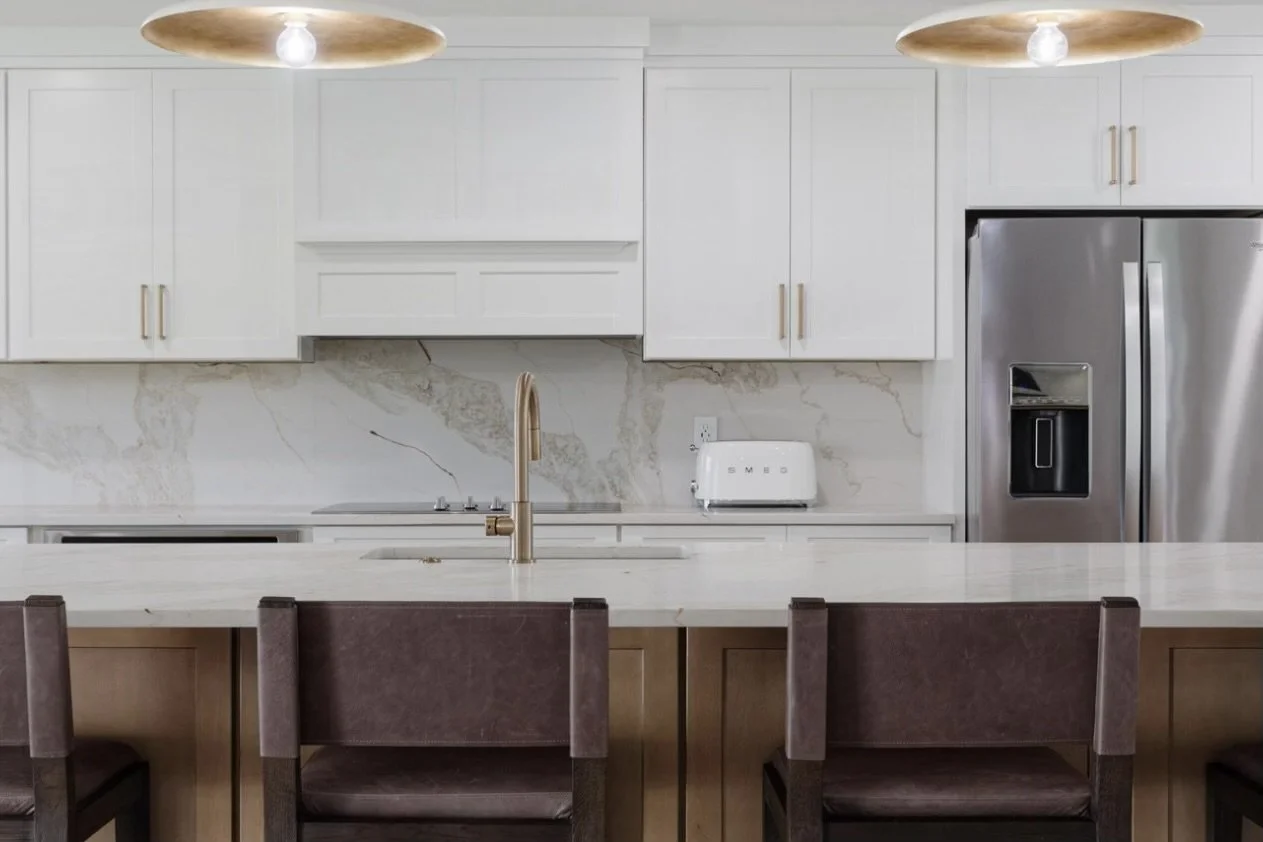 Modern kitchen with white upper cabinets, marble backsplash, stainless steel refrigerator, white countertop with a sink and gold faucet, and three dark brown chairs at a breakfast bar with wooden accents.