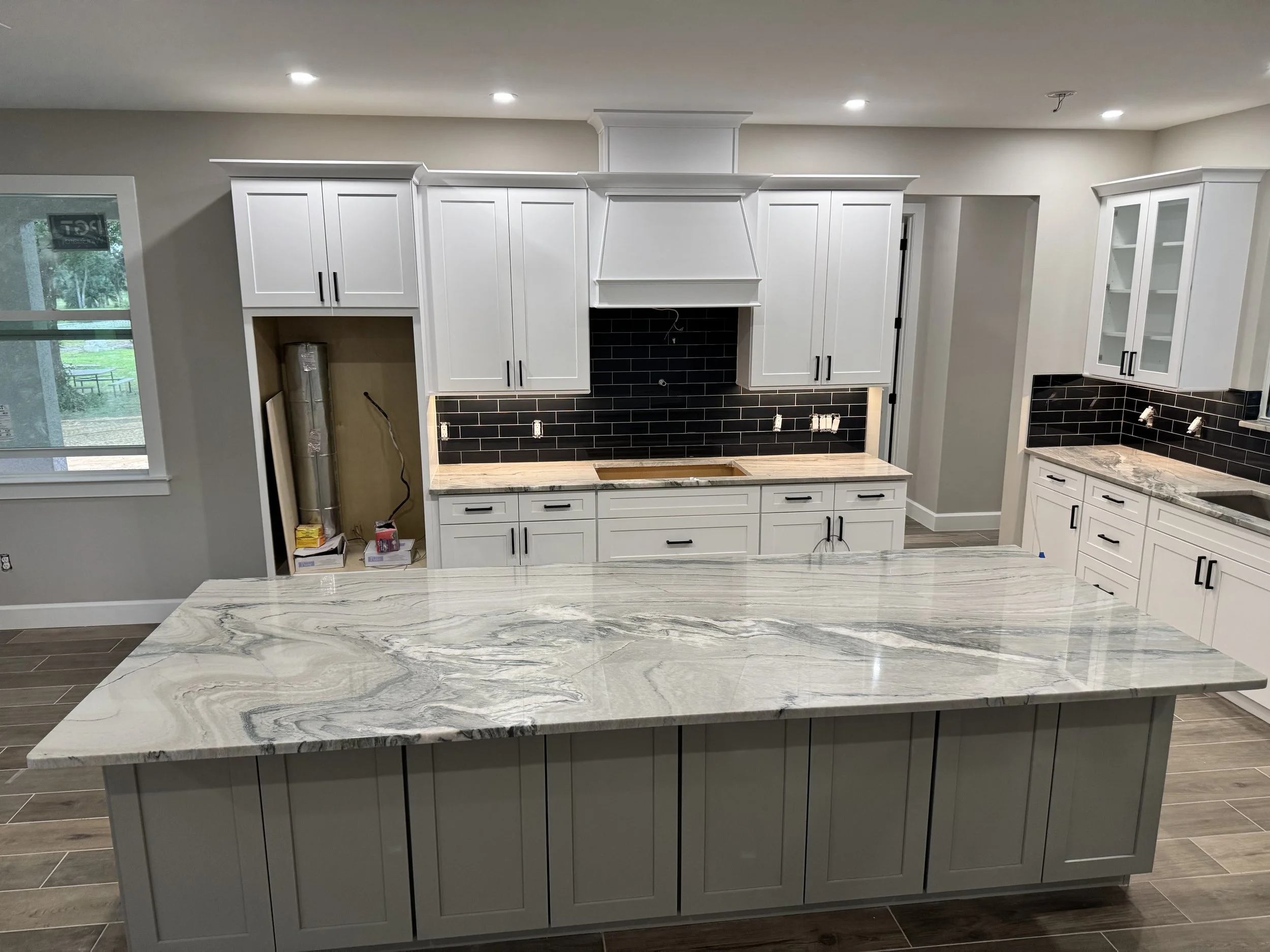 Modern kitchen with white cabinets, black backsplash, marble island, and partially visible window showing outdoor greenery.