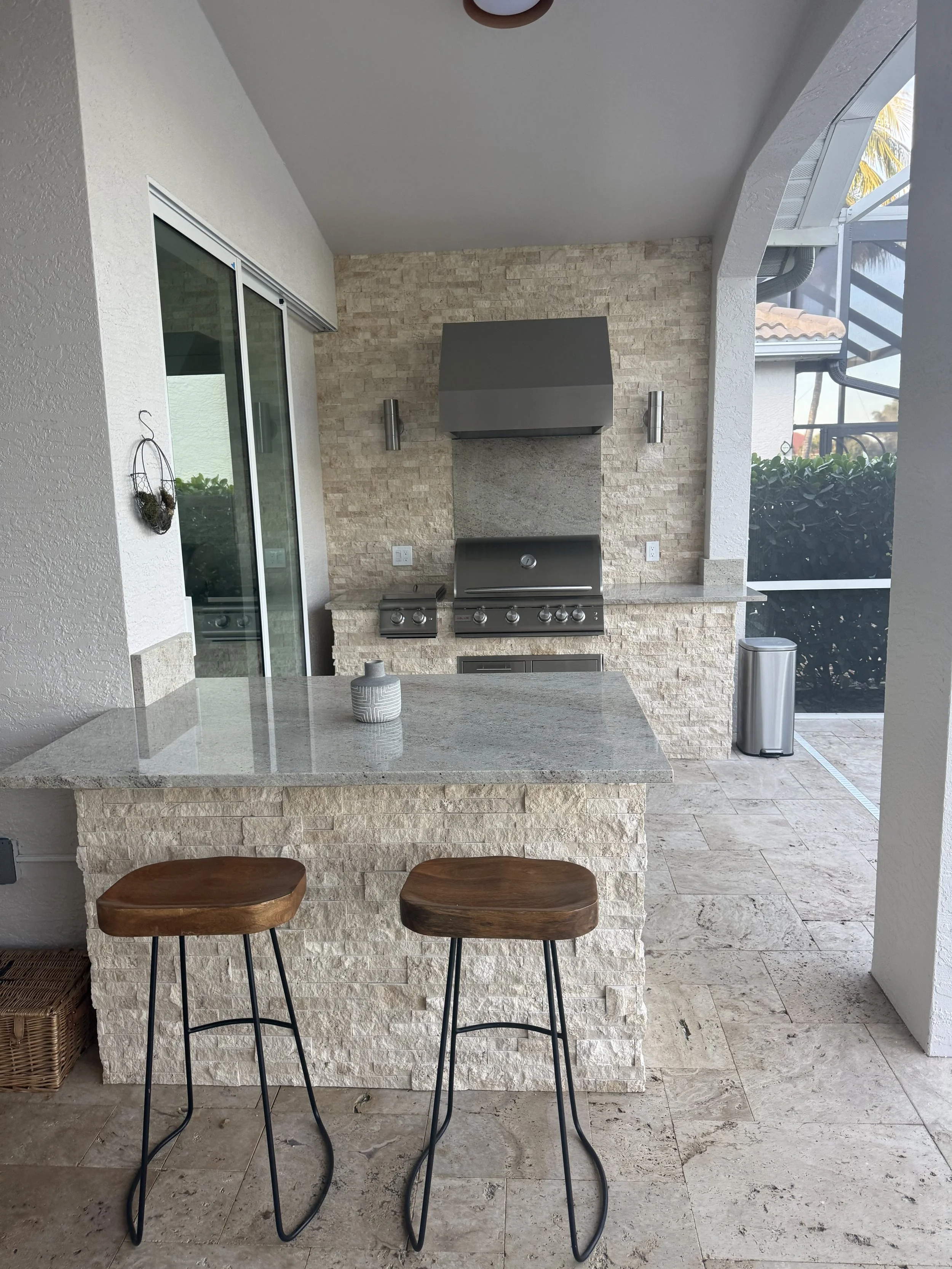 Outdoor kitchen area with a stone countertop bar and two wooden bar stools, built-in stainless steel gas grill, brick wall backsplash, and sliding glass door.