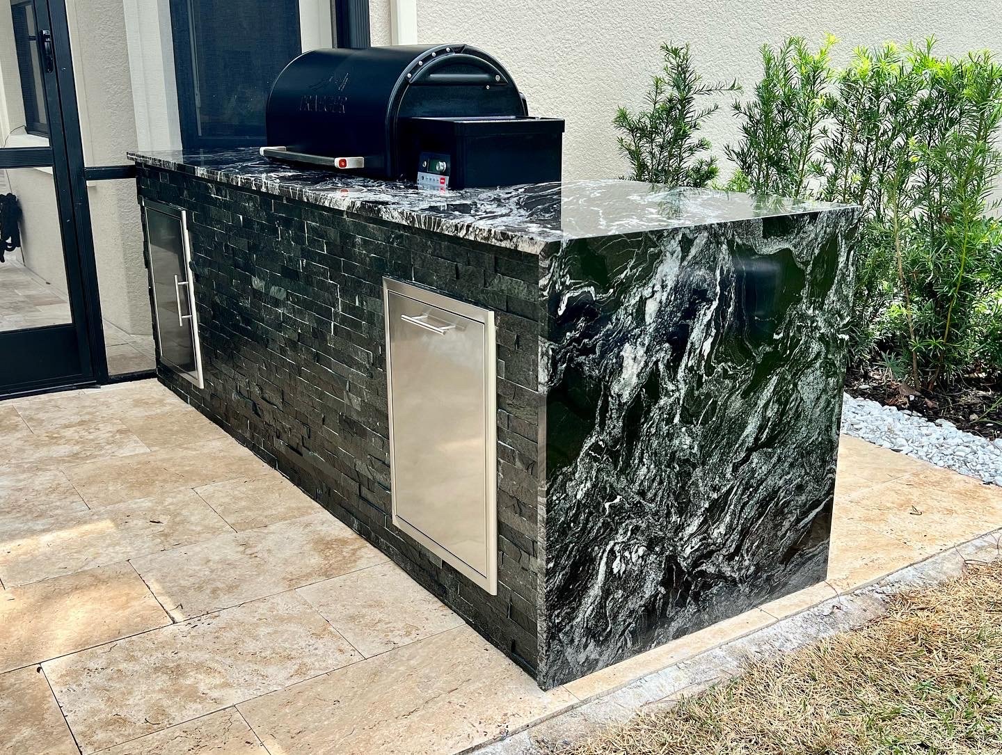 Outdoor kitchen station with a black marble countertop and decorative stone wall, equipped with a grill, cabinets, and a small appliance, surrounded by green plants.