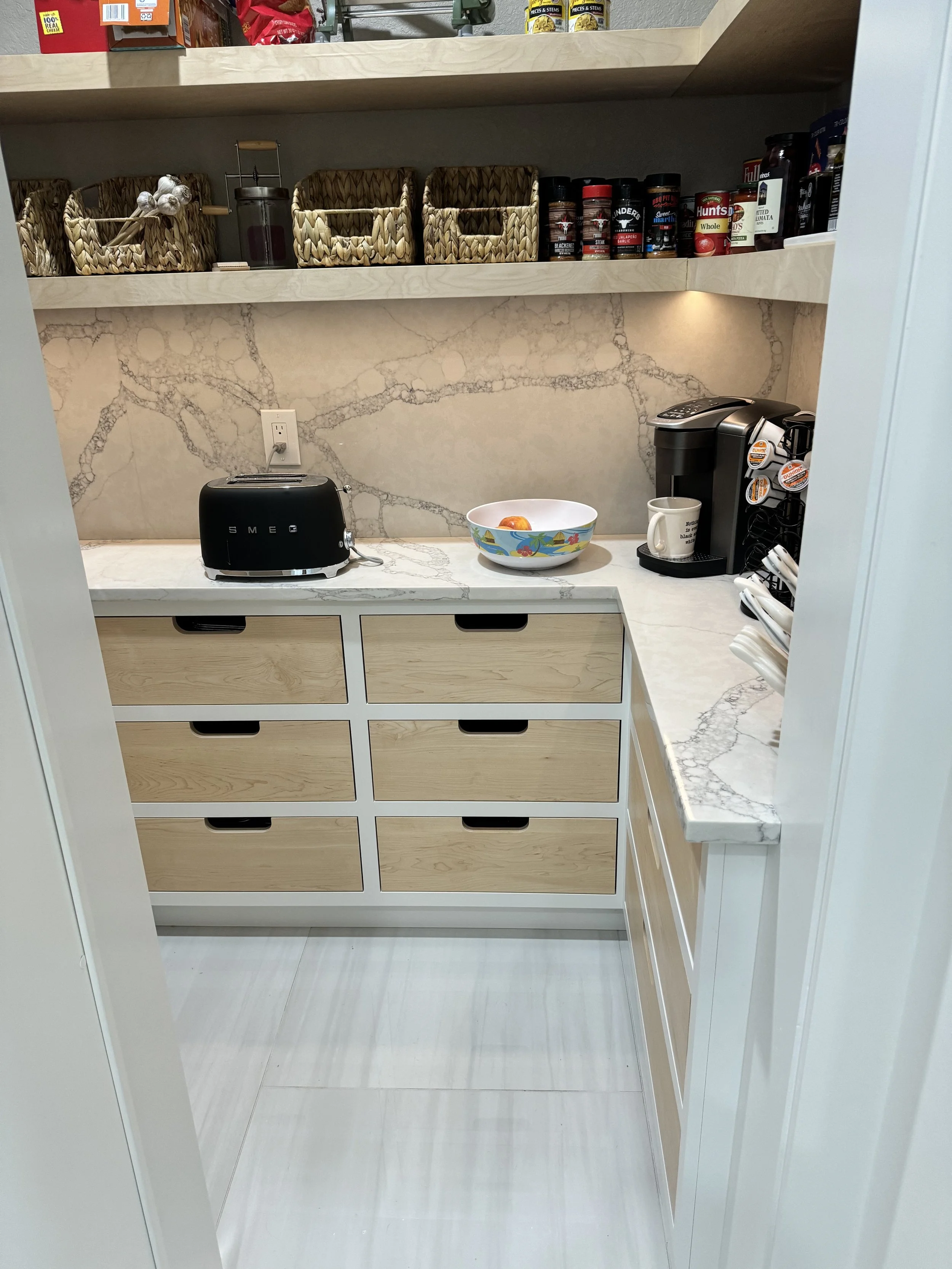 A kitchen corner with a marble countertop, beige drawers, and a backsplash with a marble pattern. On the counter, there is a black toaster, a bowl with a fruit inside, a coffee maker, and a mug. Open shelves above hold baskets, jars, spices, and groc