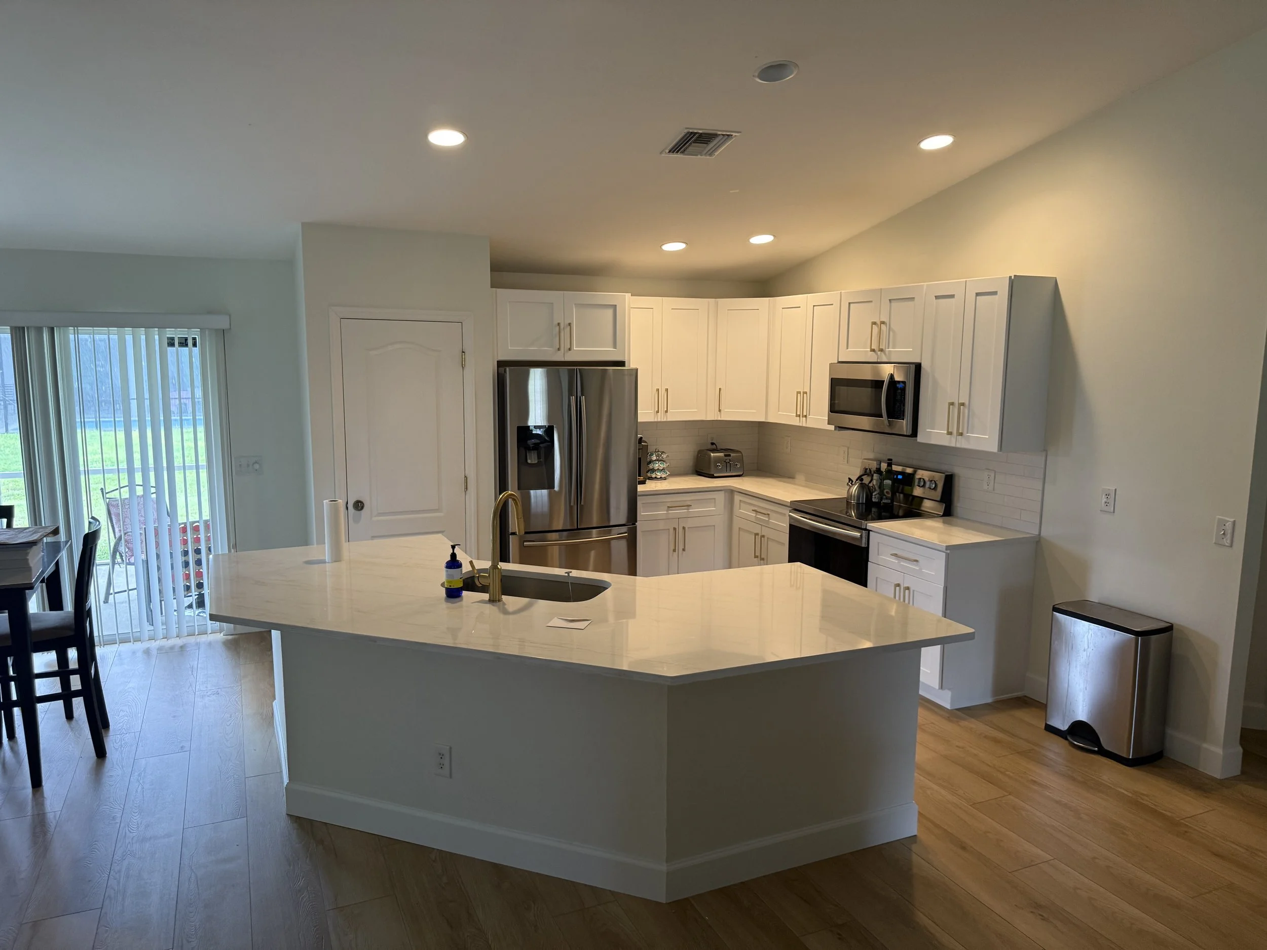 Modern kitchen with white cabinets, stainless steel refrigerator, microwave, black stove, and a large white kitchen island with a gold faucet. Sliding glass door leads to outdoor patio.
