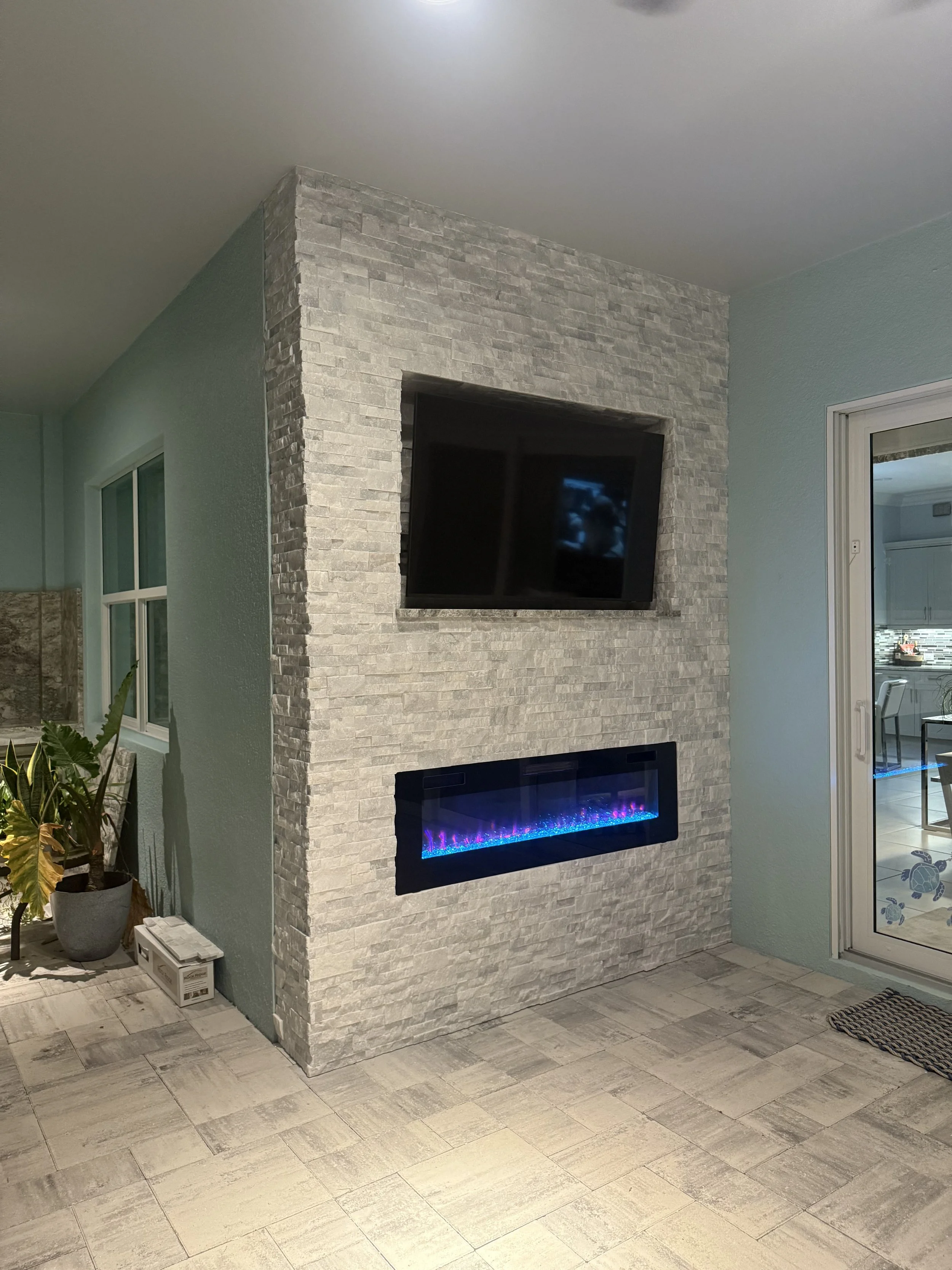 A corner of a living room with a modern stone electric fireplace and a wall-mounted flat-screen TV above it. To the left, indoor plants are visible, and to the right, a glass sliding door leading to an outdoor area can be seen.