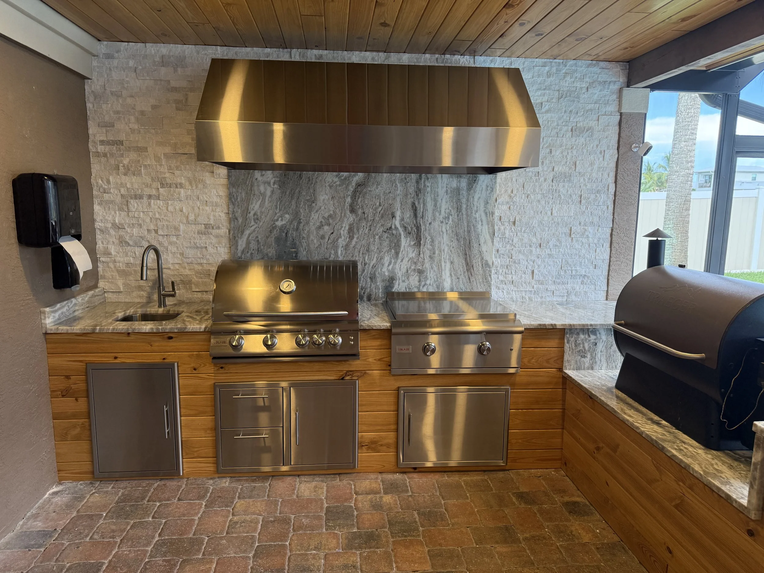 A kitchen area with a wooden and concrete backsplash, stainless steel grill, countertop, and a window showing trees outside.