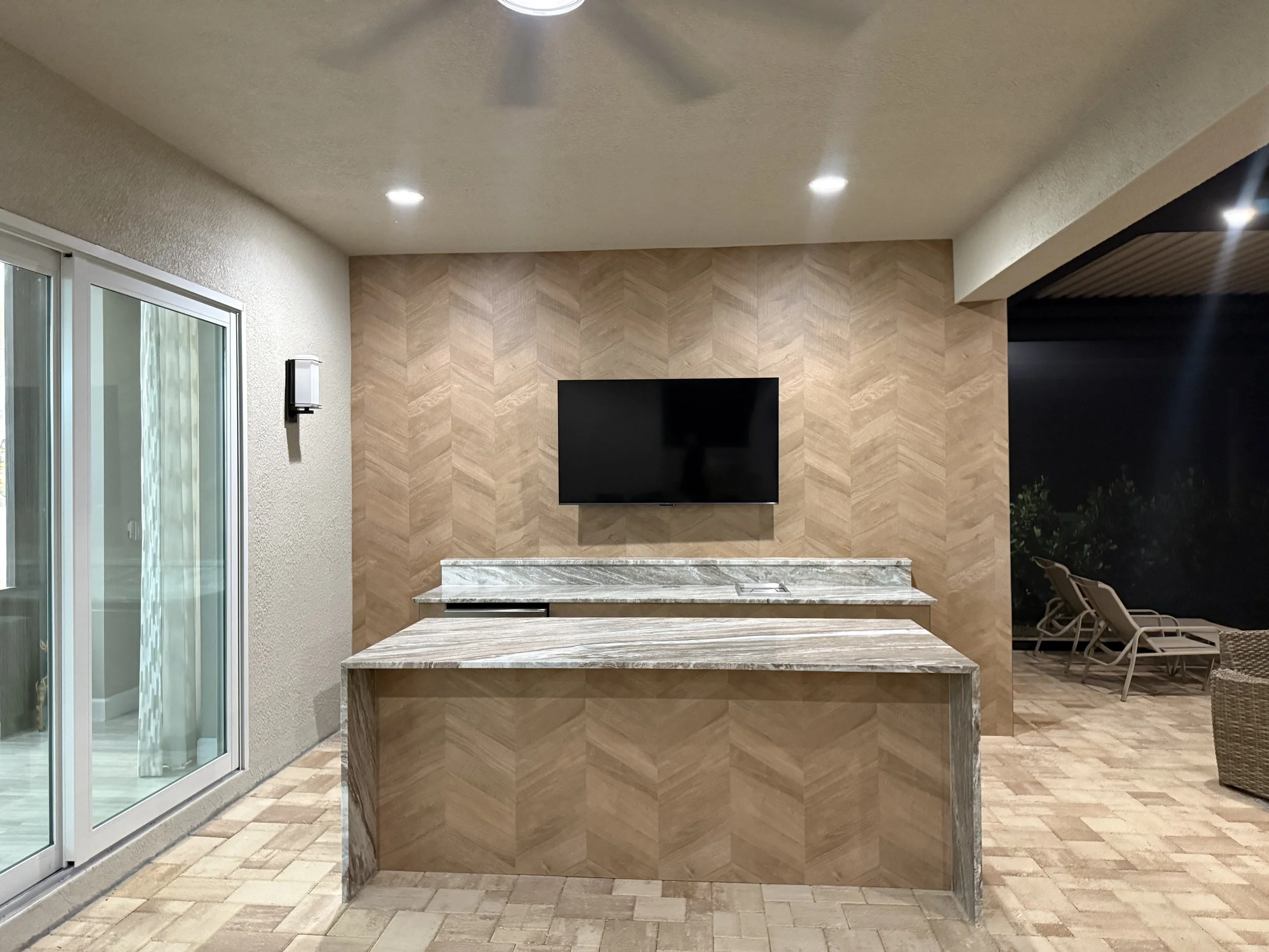 A kitchen counter with a marble surface and a herringbone patterned wood tile backsplash, mounted on a wall with a television, with sliding glass doors on the left and outdoor chairs on the right.