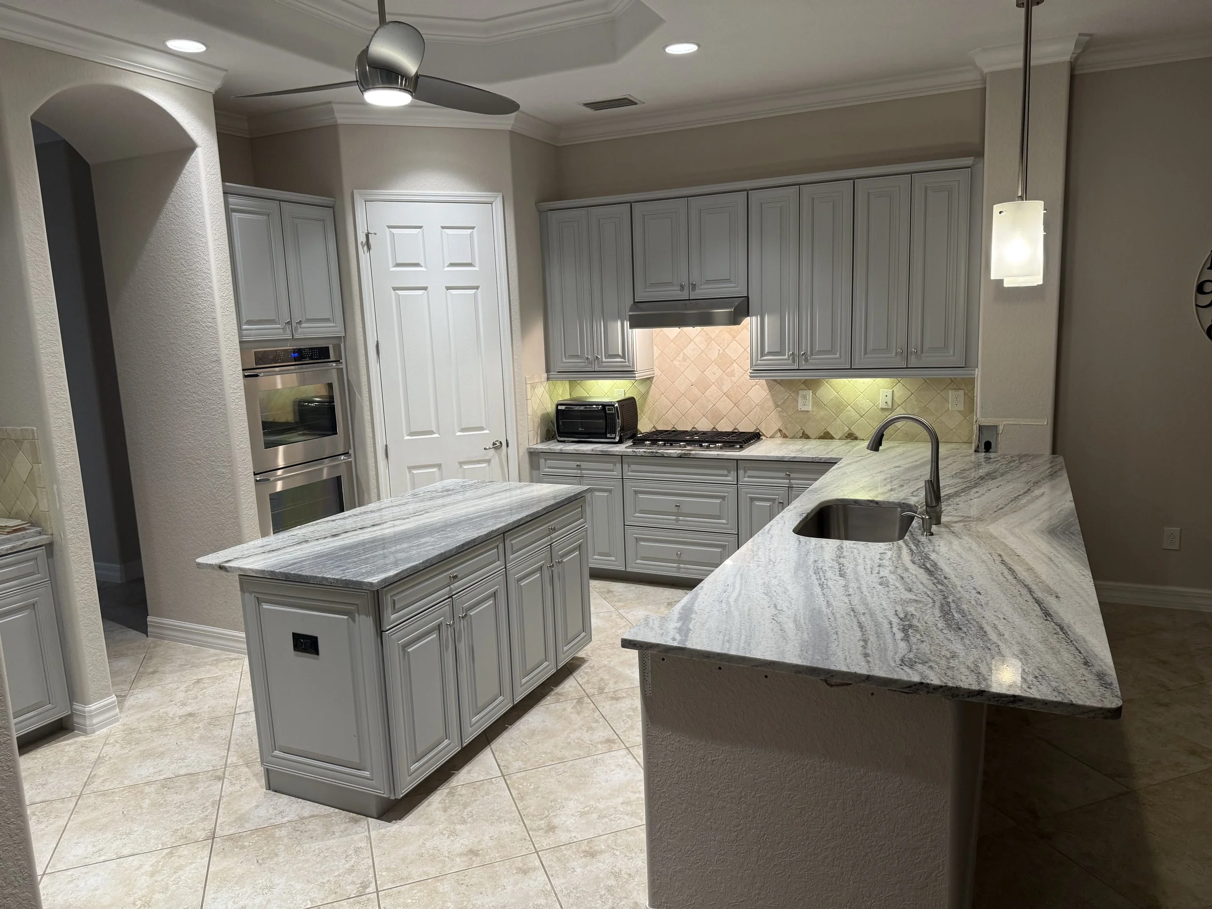 Modern kitchen with white cabinets, marble countertops, beige tile backsplash, stainless steel oven, microwave, and vent hood, and a ceiling fan.