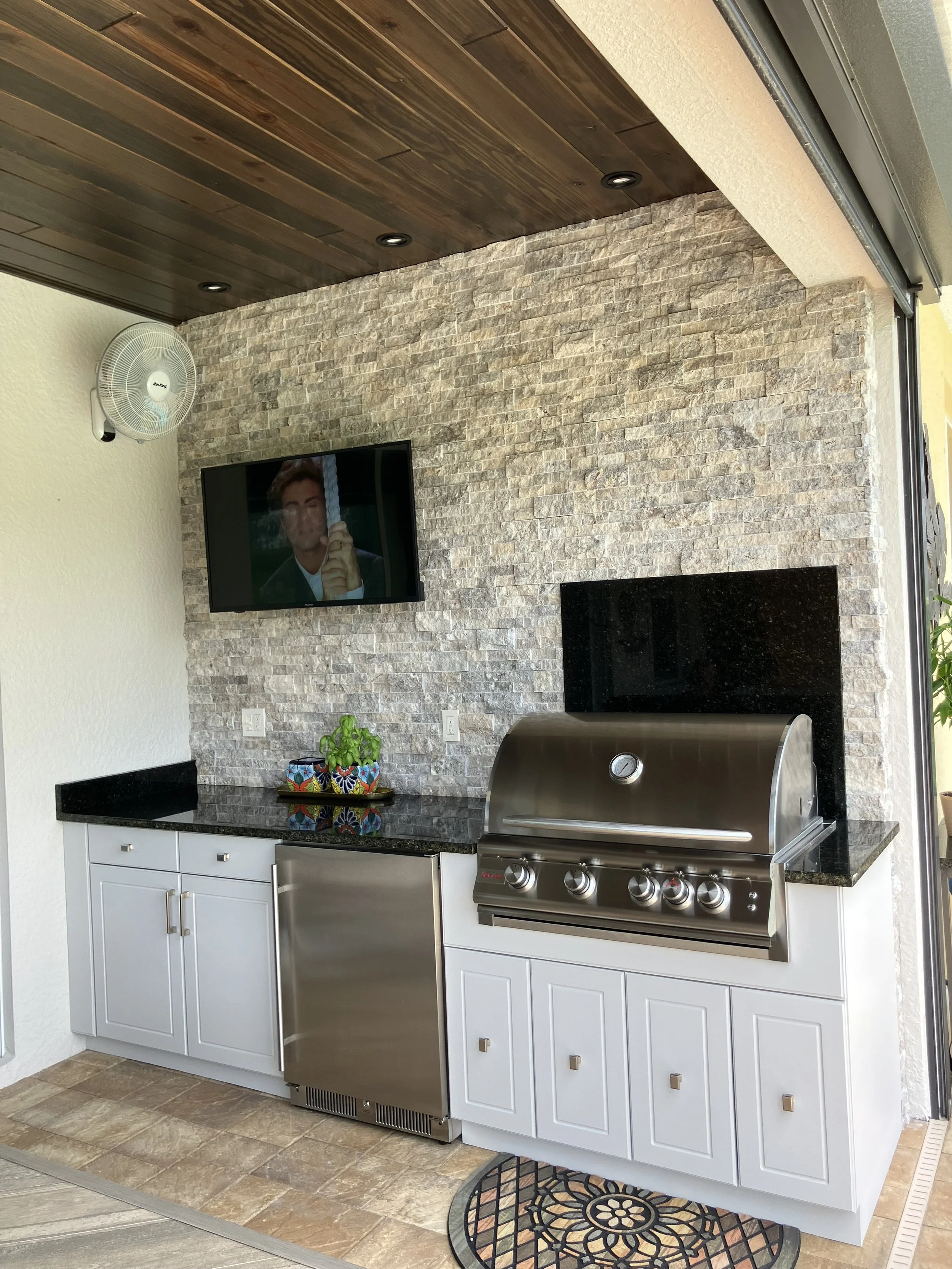 Outdoor kitchen with a stone wall, a stainless steel grill, white cabinets, a black granite countertop, a wall-mounted flat-screen TV, a ceiling with wooden panels and recessed lighting, a small fan on the wall, a colorful potted plant, and a decorat