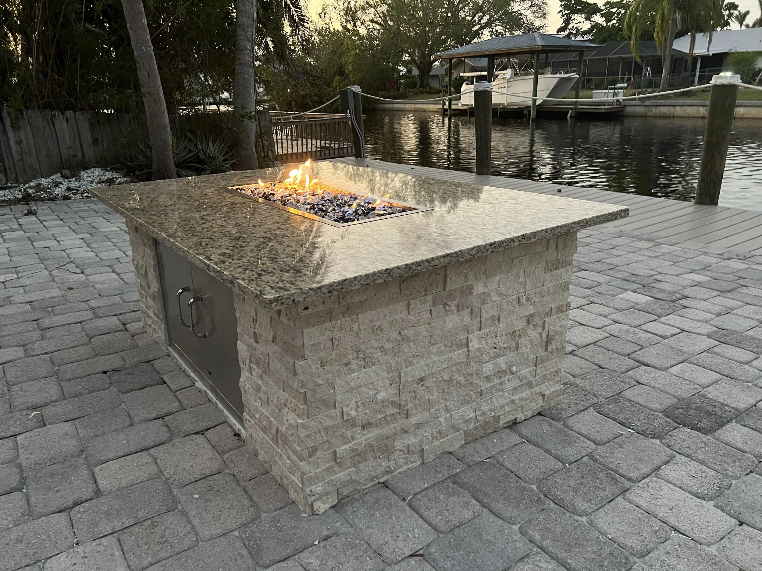 A backyard with a fire pit on a stone patio overlooking a canal. There are boats docked along the water and trees surrounding the area during dusk.