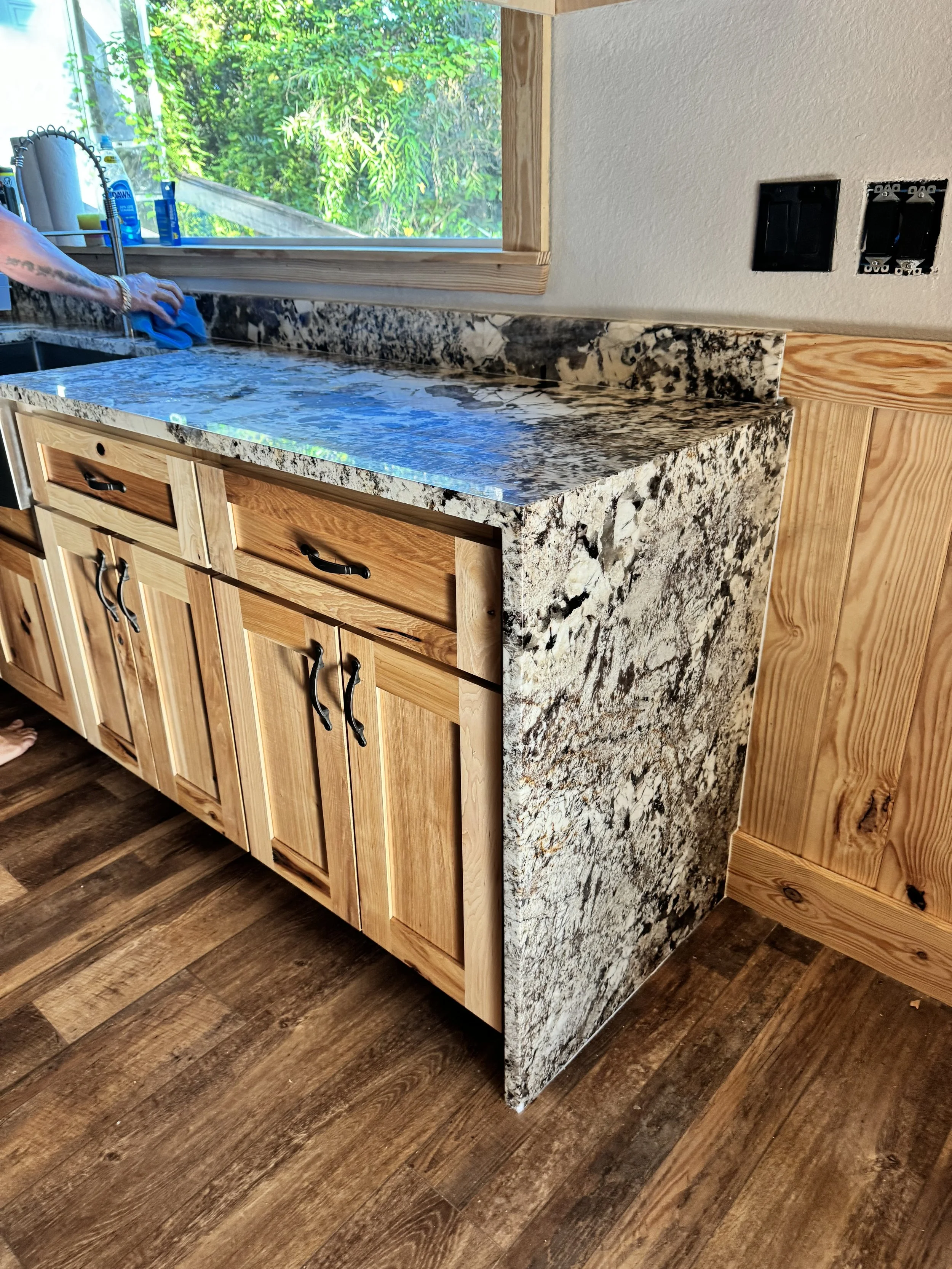 A kitchen corner with a granite countertop, wooden cabinets, and a window overlooking greenery.