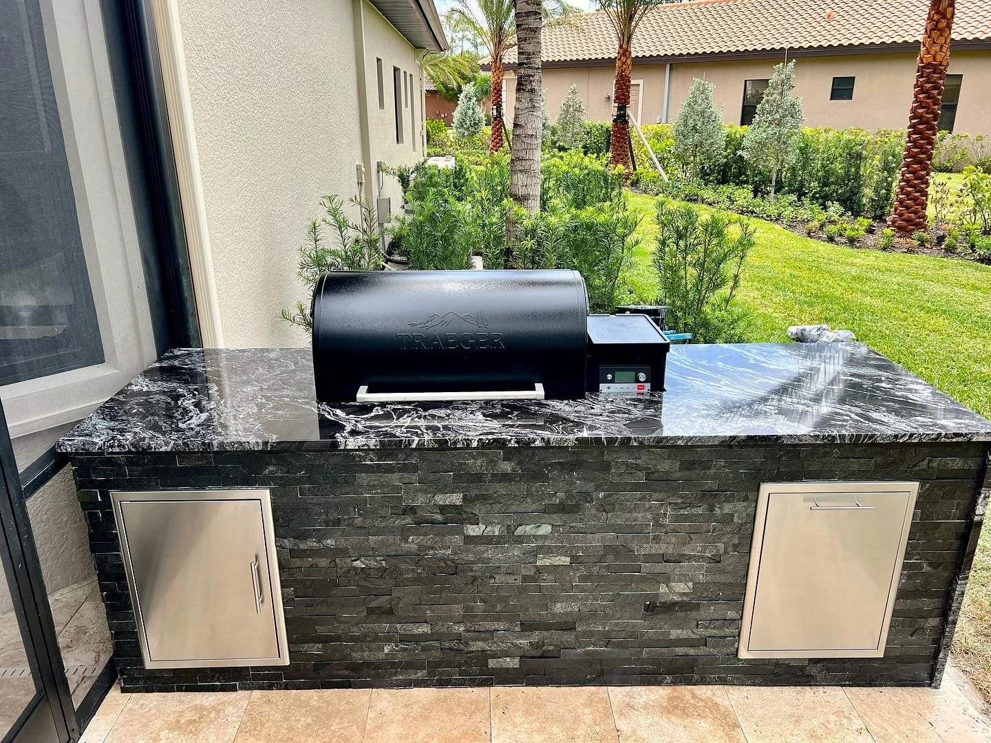 Outdoor kitchen counter with black marble surface, a black Traeger pellet grill, silver cabinet doors, and a backyard with green plants, palm trees, and a beige house in the background.