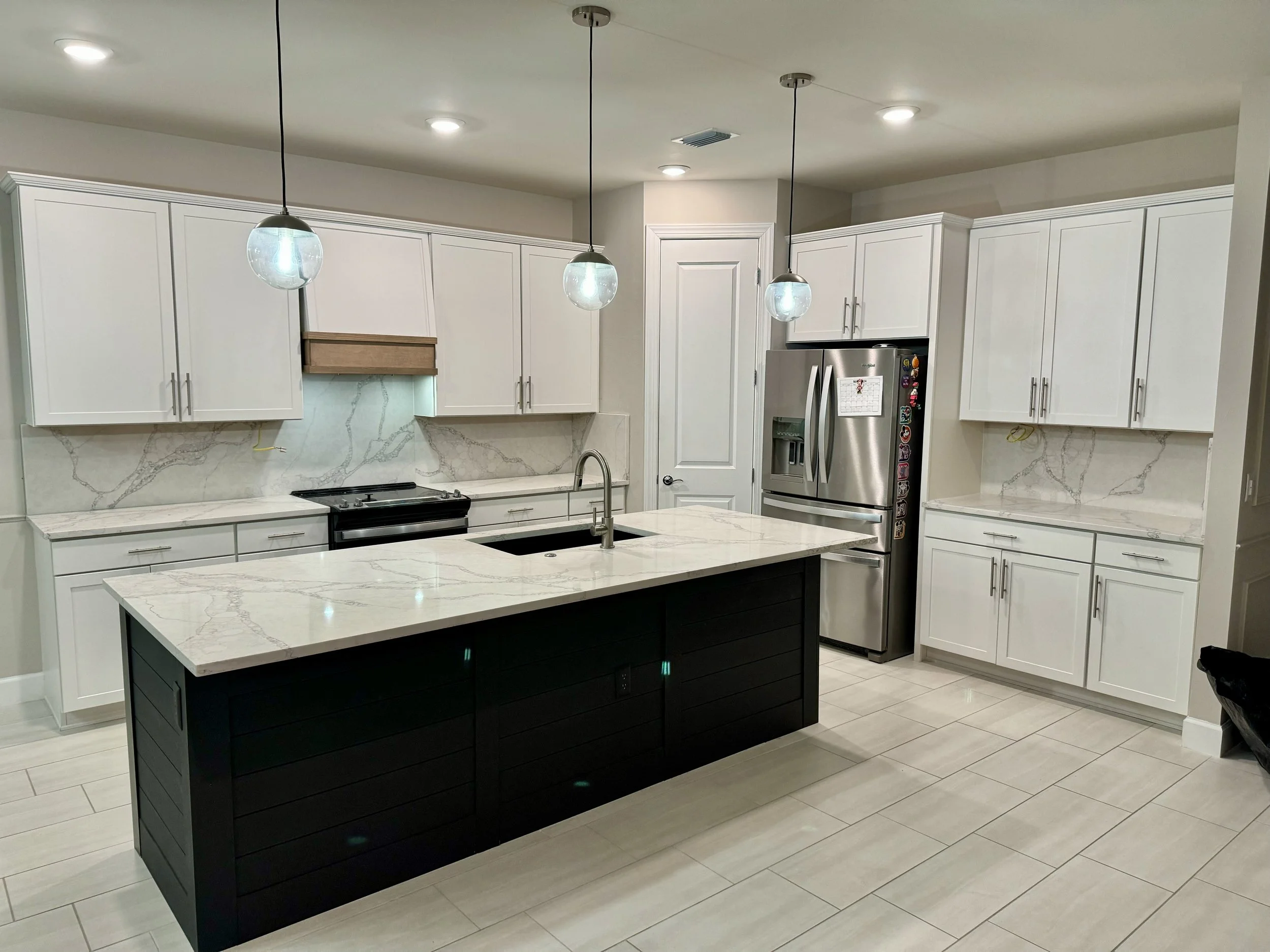 Modern kitchen with white cabinets, marble countertops, a black kitchen island, pendant lights, a stainless steel refrigerator, and beige tile flooring.