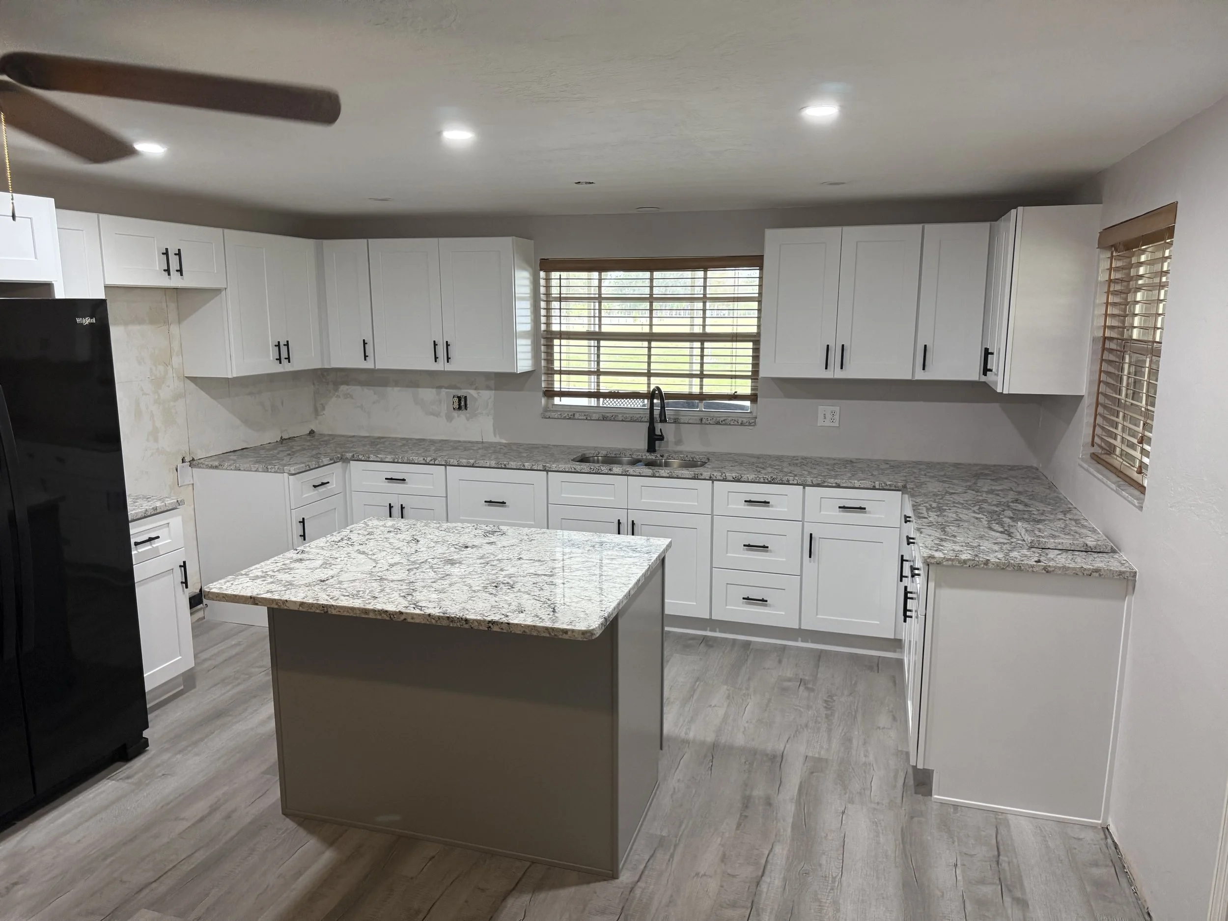 Modern kitchen with white cabinets, granite countertops, a kitchen island, black handles, a black refrigerator, hardwood flooring, and three plastic window blinds.
