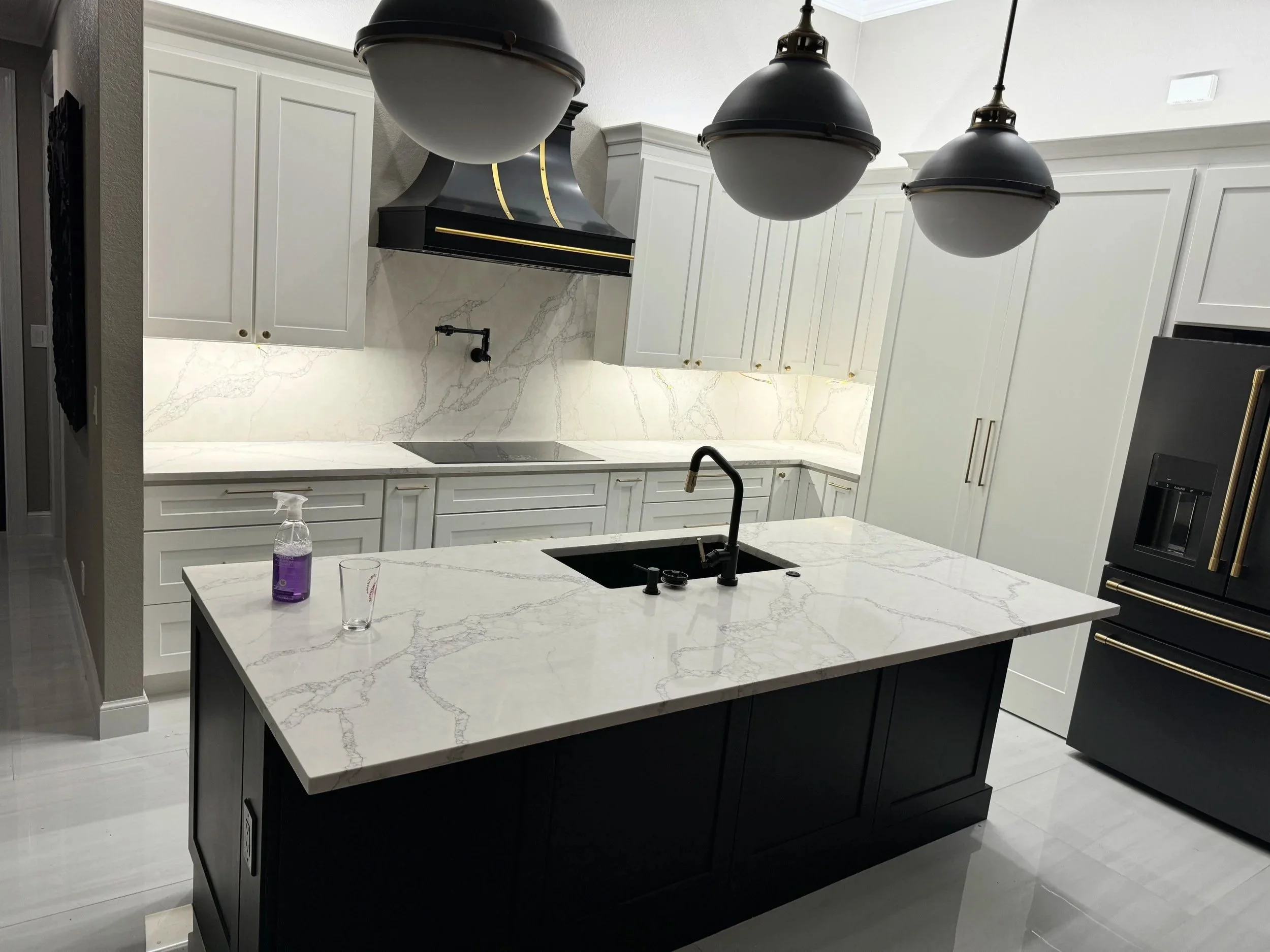 Modern kitchen with white cabinets, black and gold hood, marble countertops, and three black pendant lights hanging above a black island with a marble top. Bottled water and a glass are on the island.