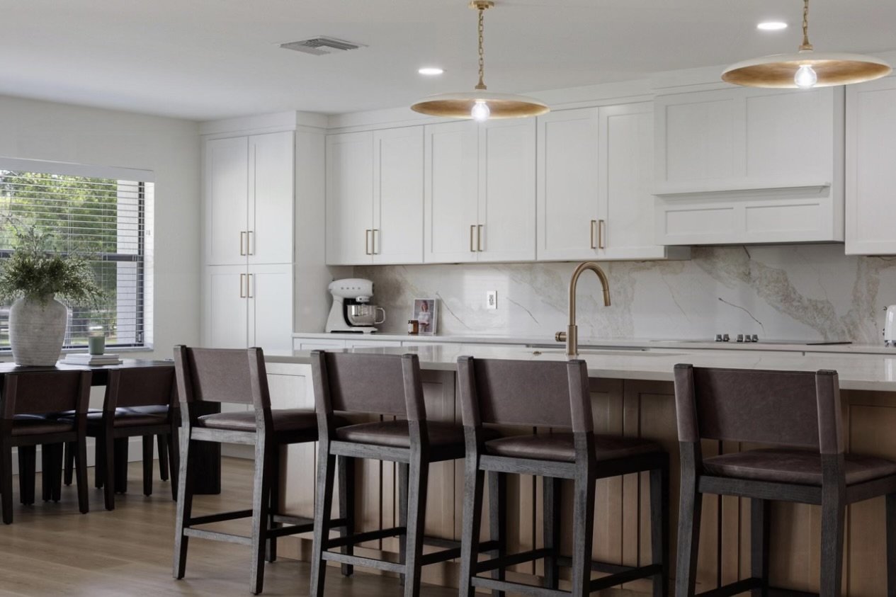 Modern kitchen with white cabinets, marble backsplash, gold fixtures, a large window with blinds, a dark wood breakfast bar with four chairs, a potted plant, and a coffee maker.