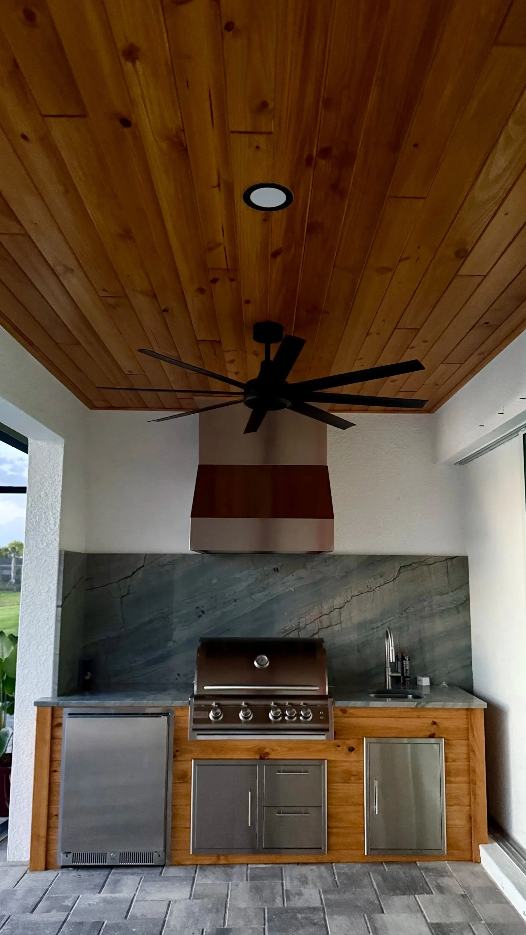 An outdoor kitchen area featuring a wooden ceiling, black ceiling fan, a stainless steel grill, a modern gray cabinet, and gray tiled flooring.