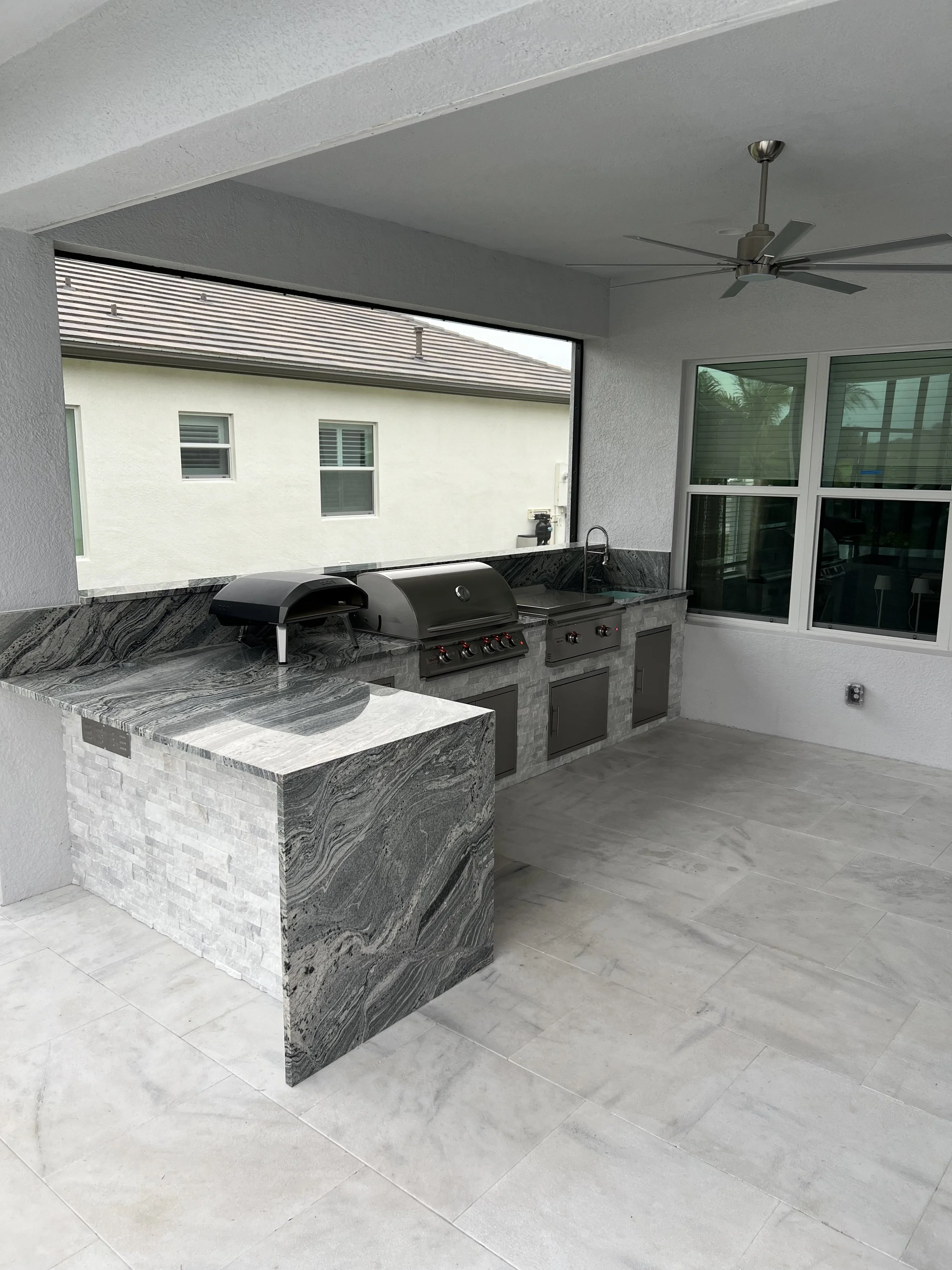 An outdoor kitchen area with a marble countertop and a grill, partially enclosed with an open window view of neighboring houses and a ceiling fan overhead.